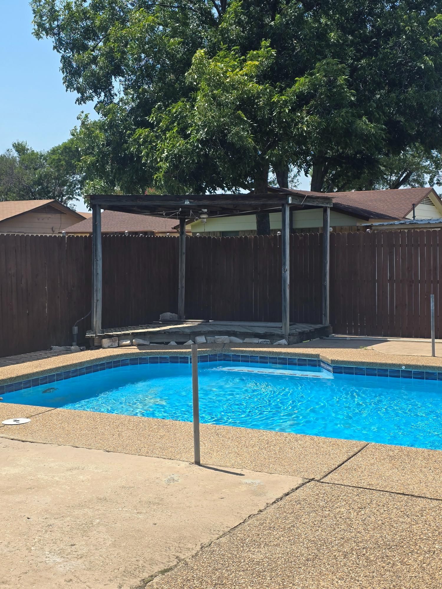 Backyard pool with a pergola structure, wooden fence, and blue water on a sunny day.
