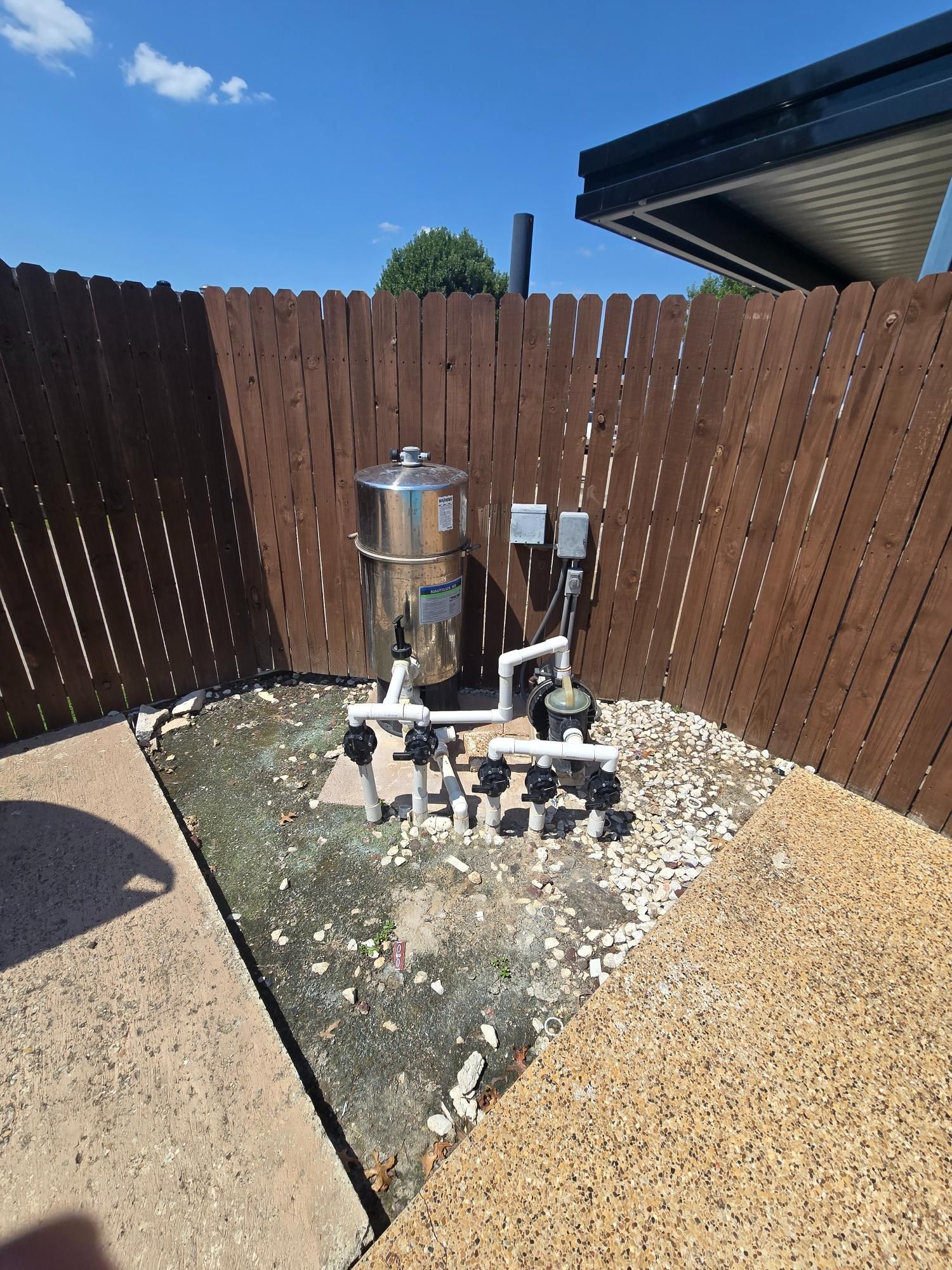 Pool filter equipment enclosed in a wooden fenced area, set against a blue sky.