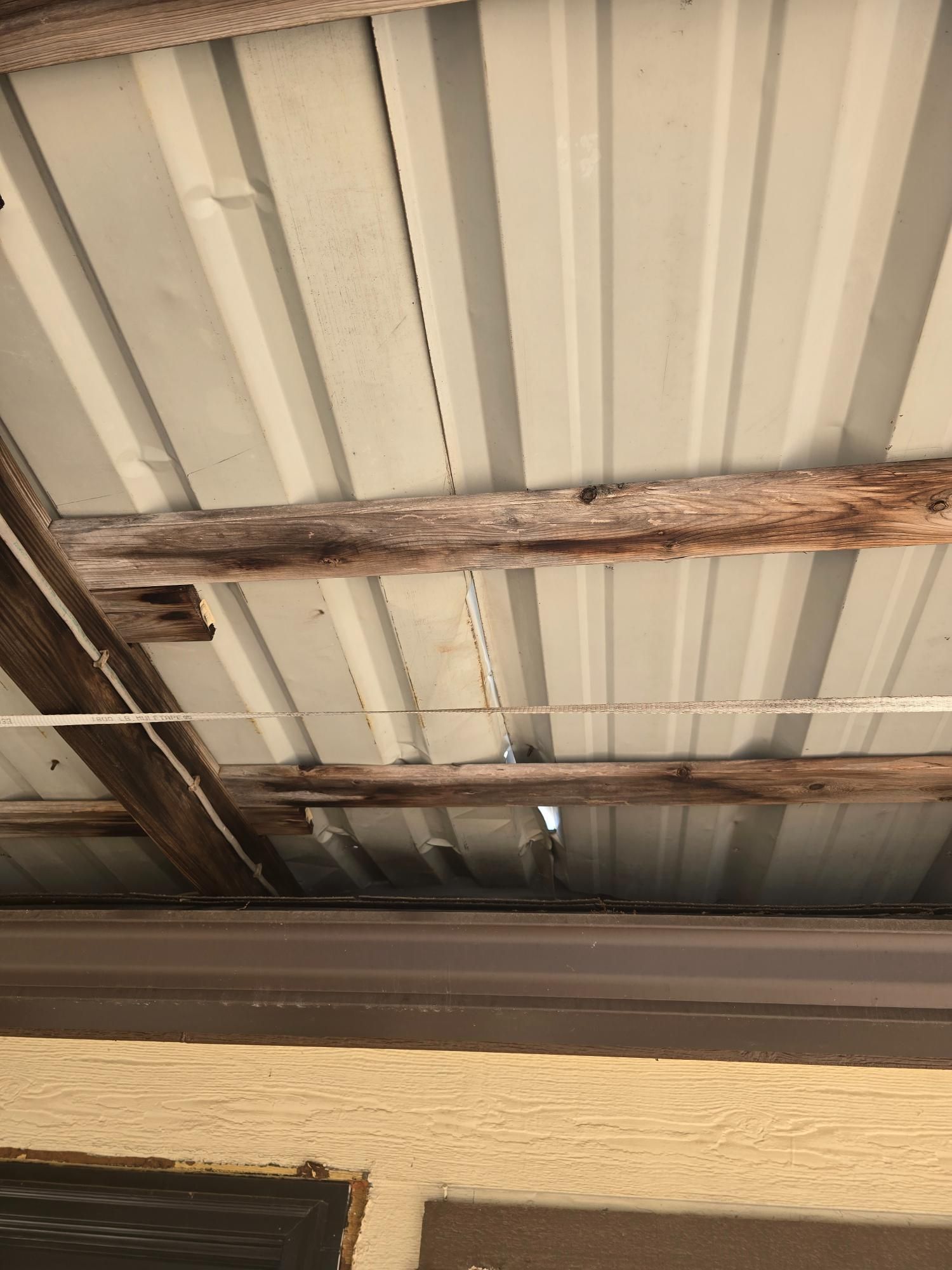 View of a corrugated metal roof supported by wooden beams, with a hole and discoloration visible.
