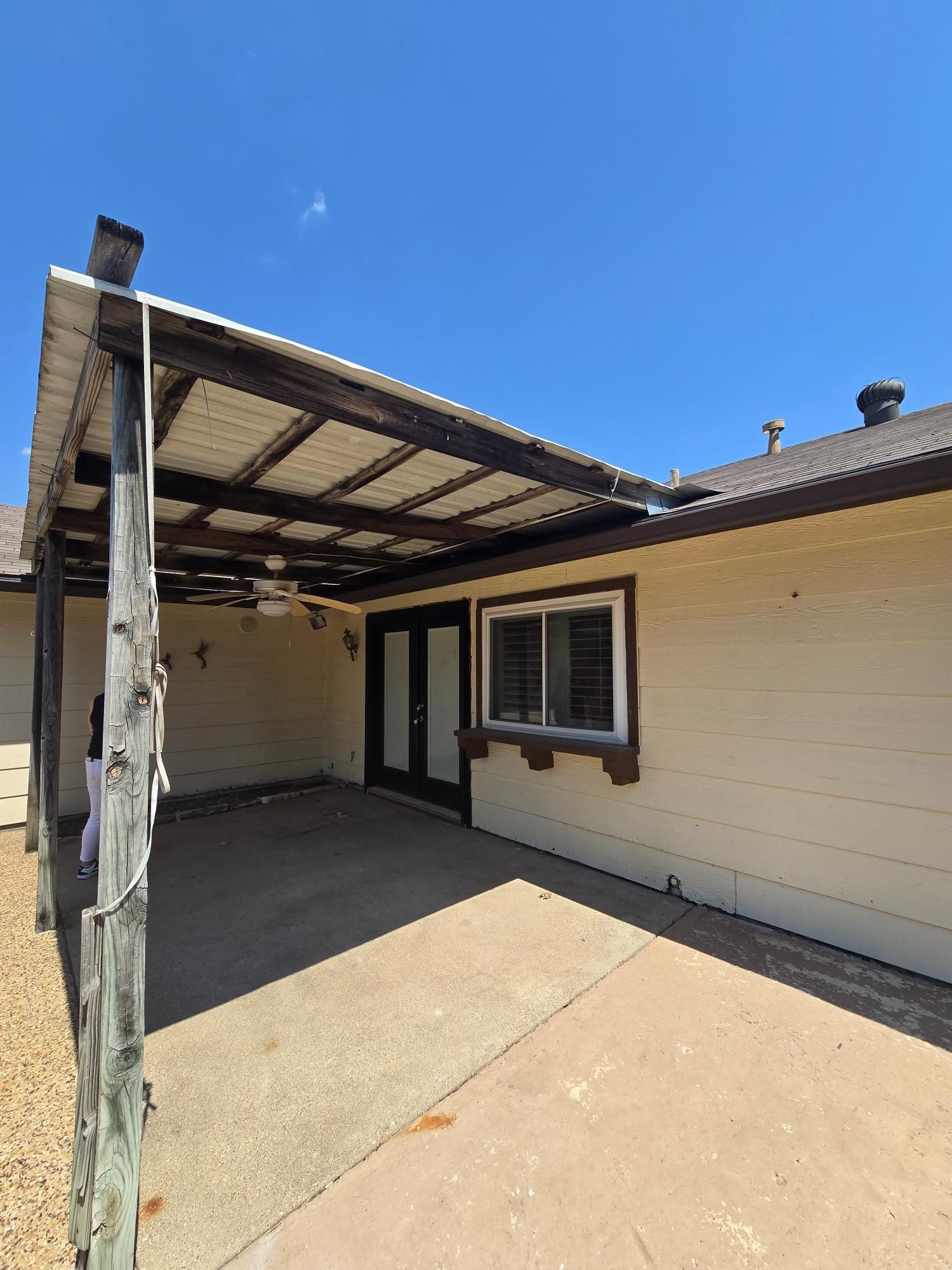 Backyard patio with wooden beams and a tan house under a blue sky.