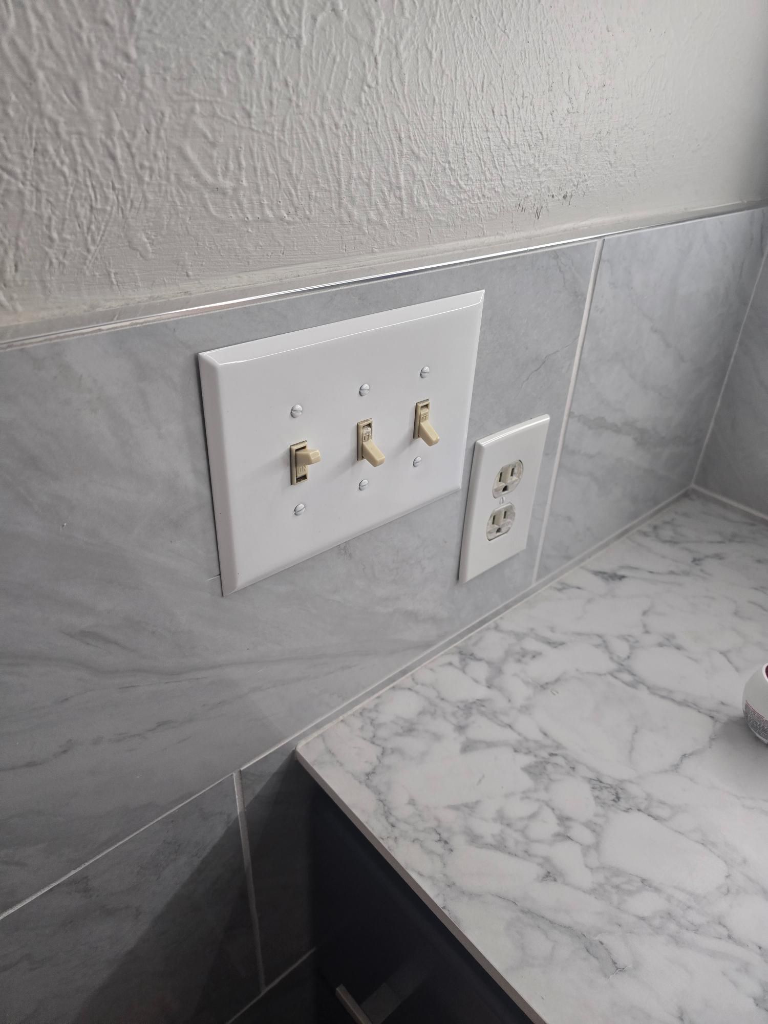 White light switches and outlet on gray tiled wall above a marble counter.
