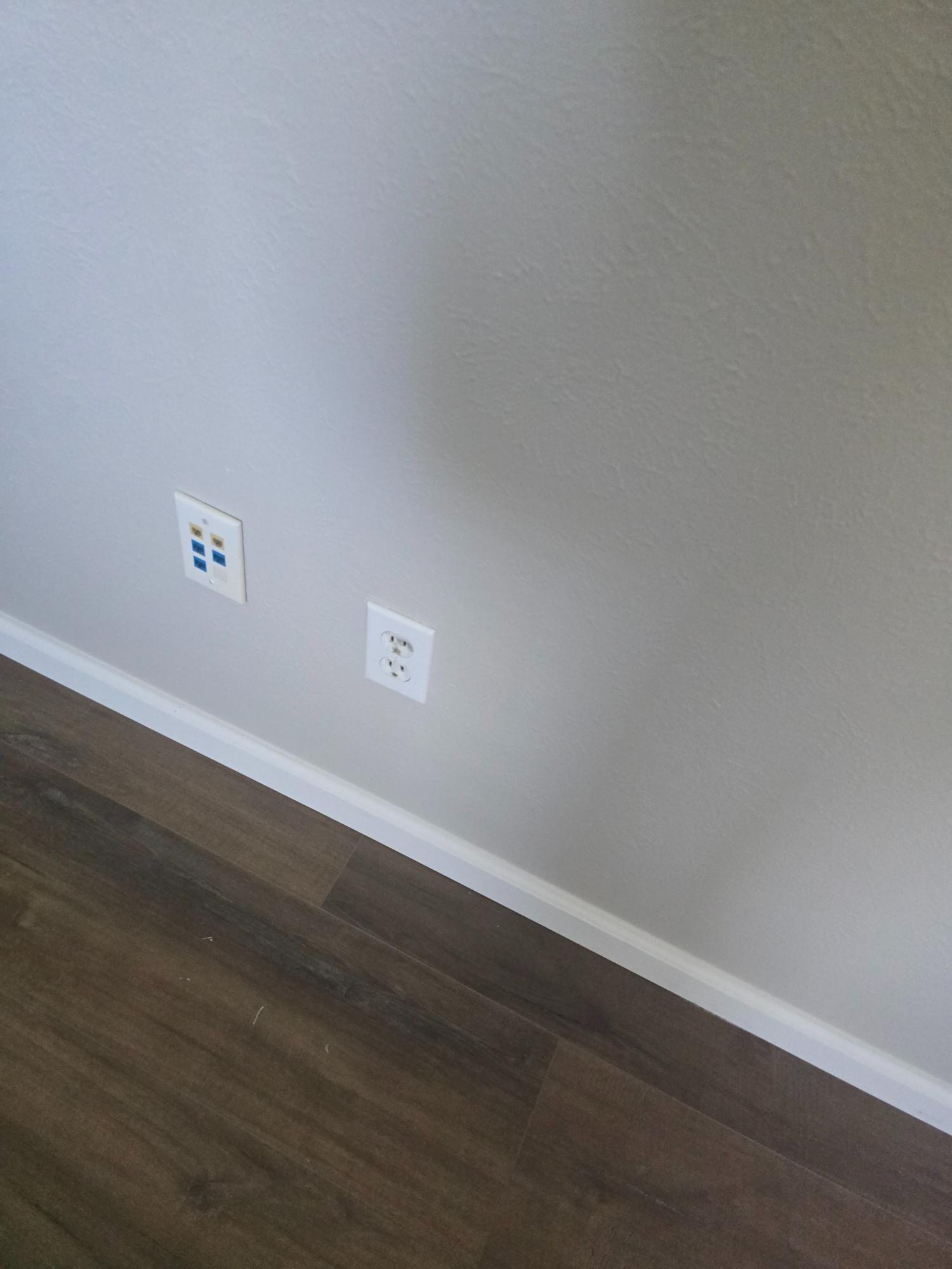 Two electrical outlets on a gray wall above dark wood flooring; one has blue covers.