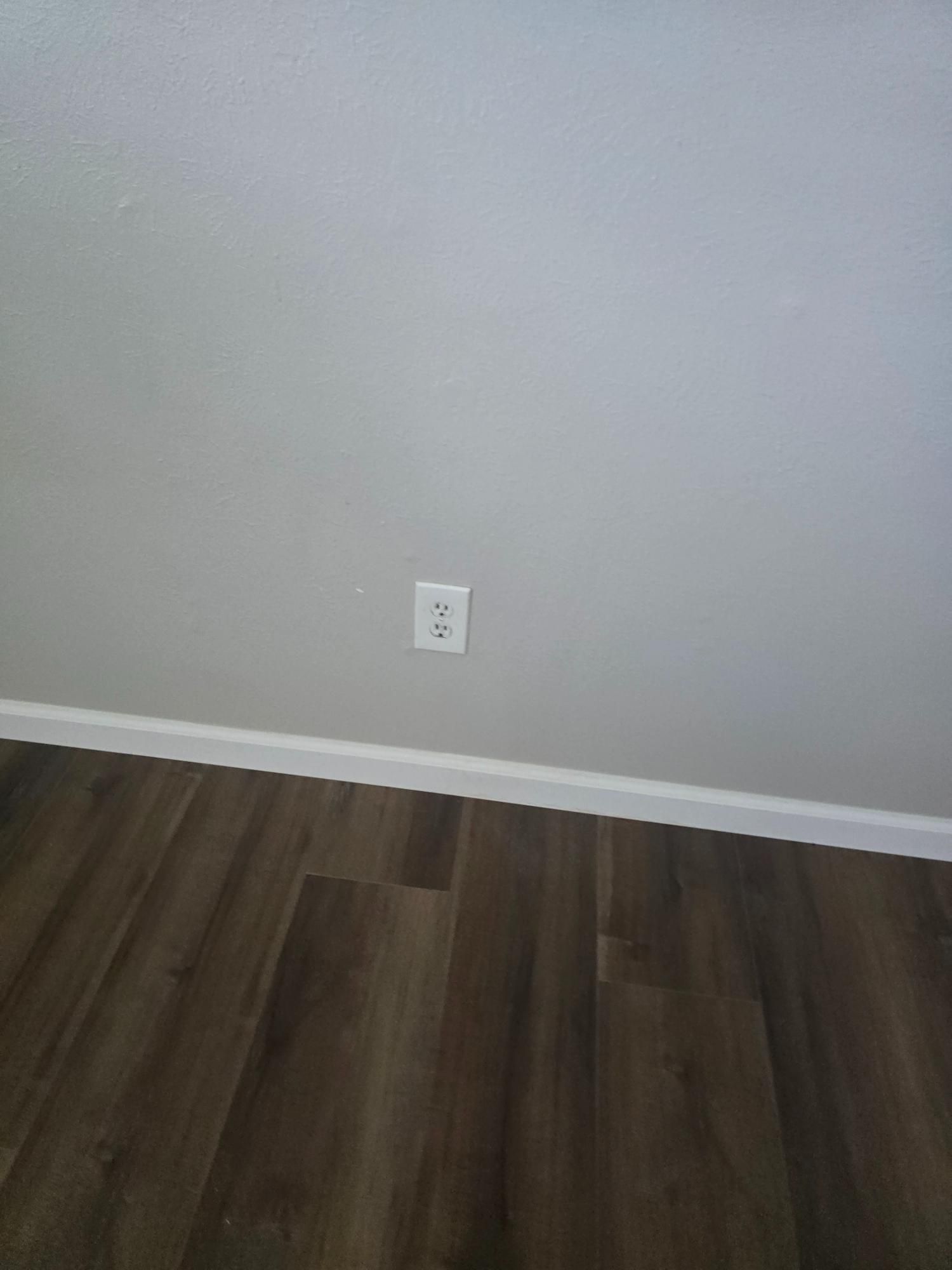 Electrical outlet on a gray wall above brown wood-look flooring and white baseboard.