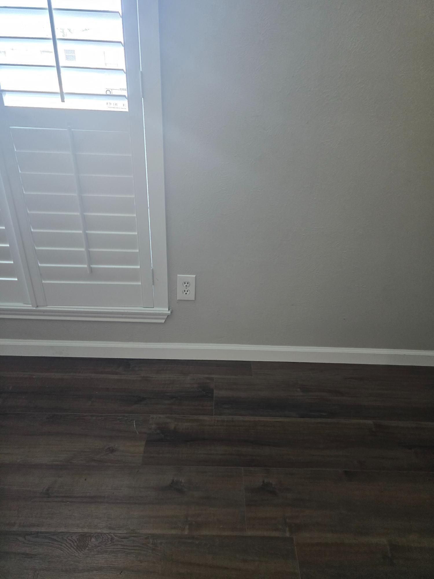 Close-up view of a room corner with a window, outlet, and wood-look flooring.