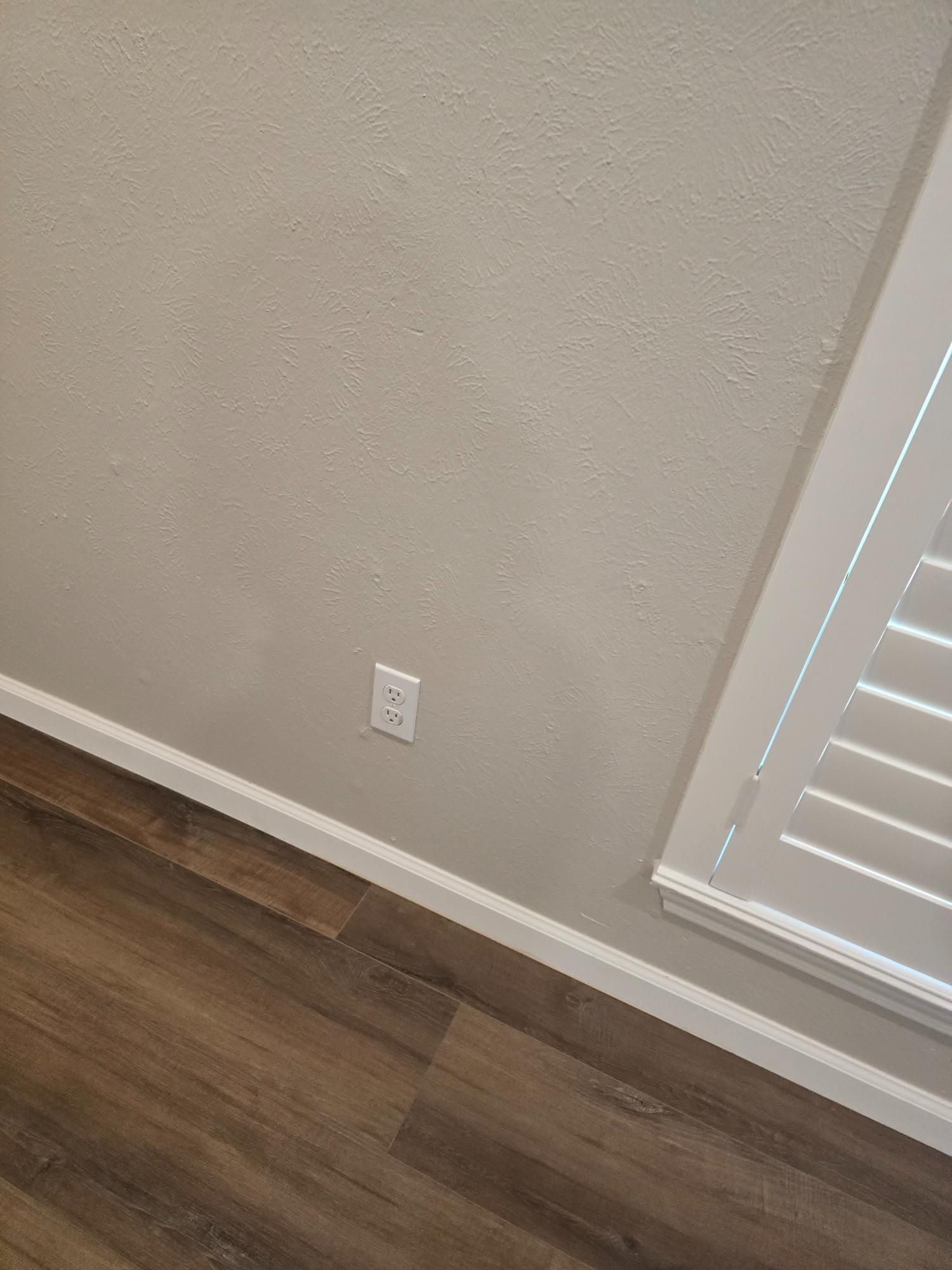 Beige wall with outlet, white trim and shutters, and brown wood-look floor.