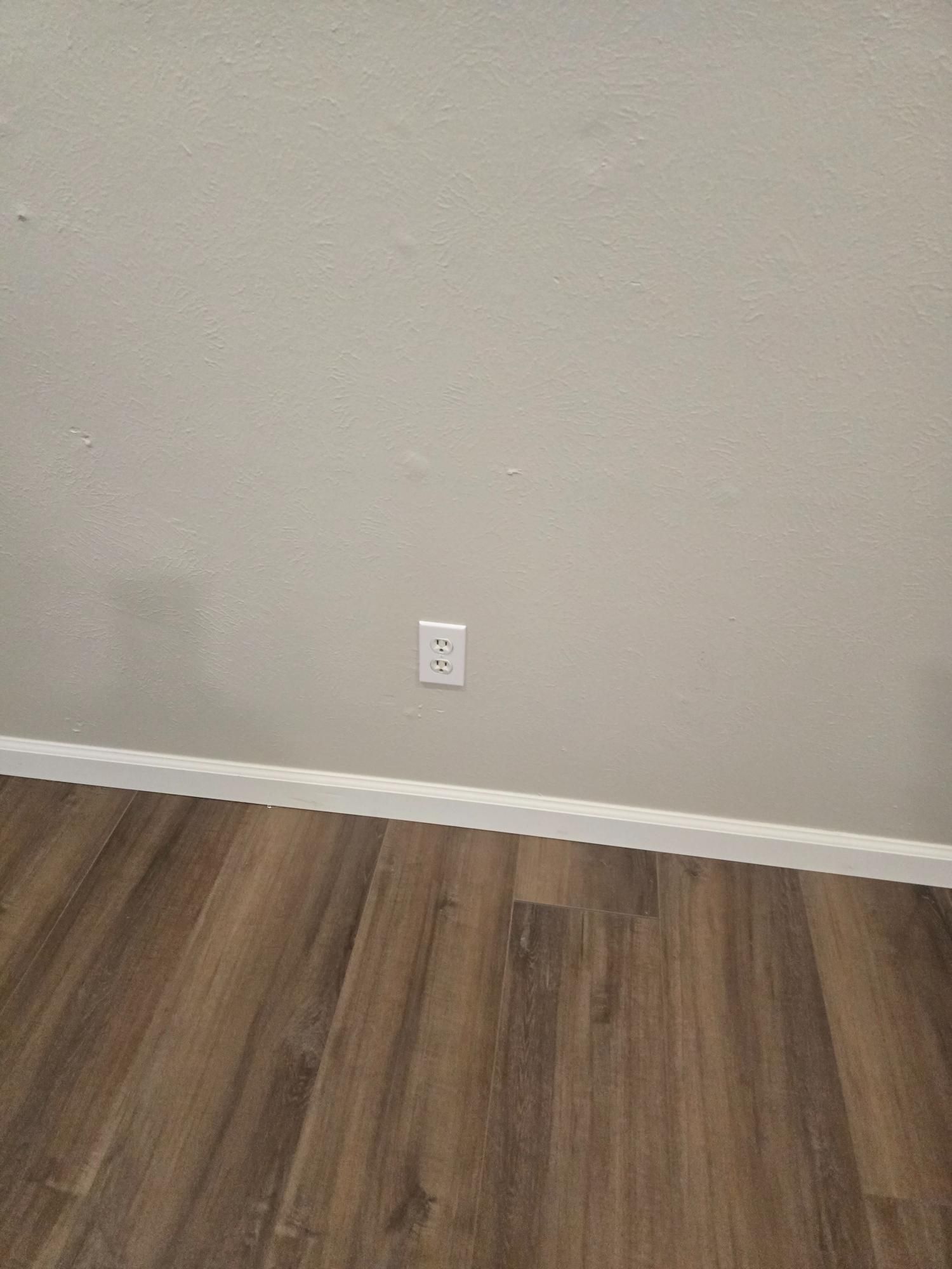 Wall with white electrical outlet and brown wood-look flooring.