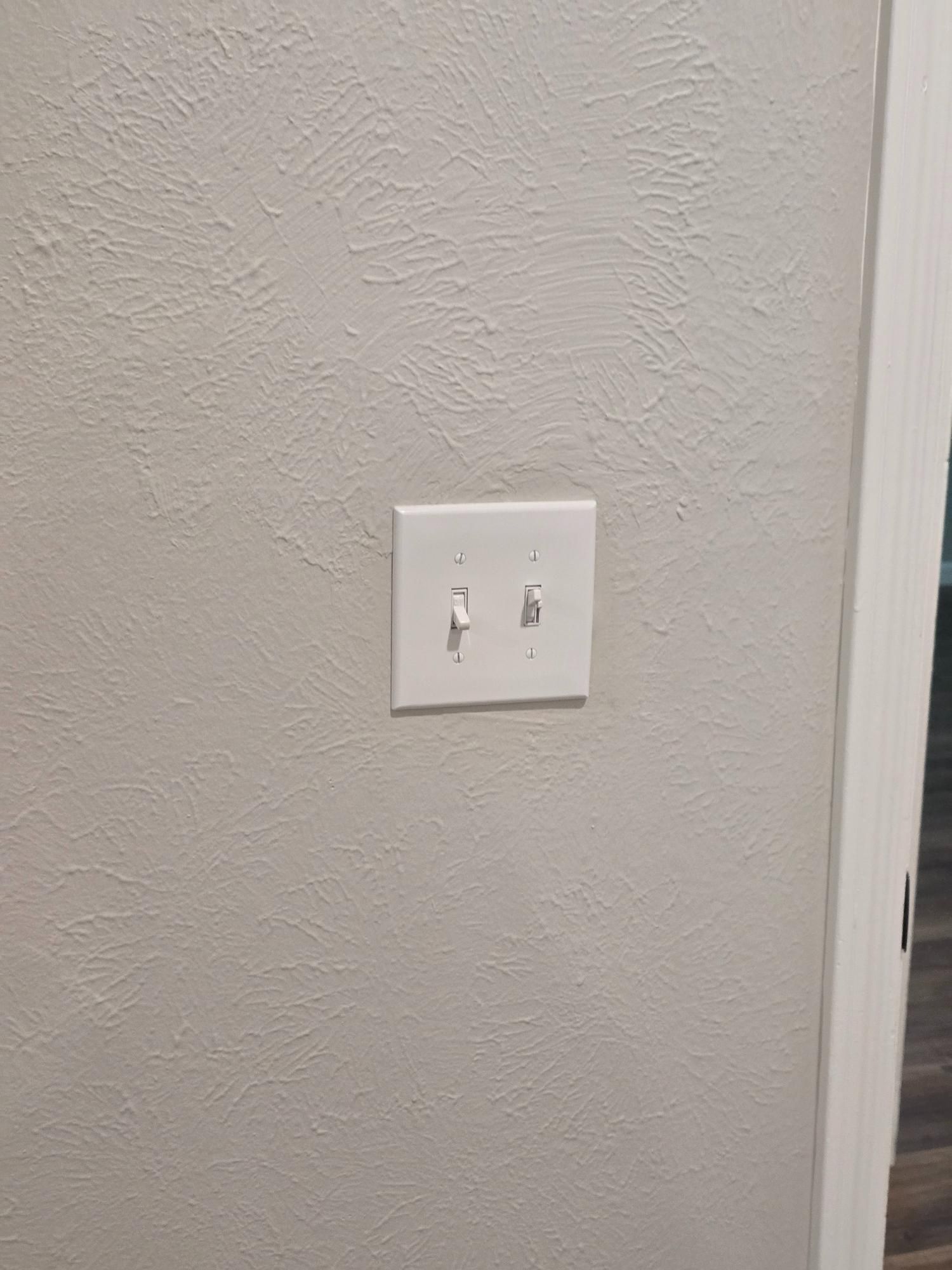 Two white light switches on a textured, off-white wall next to a door frame.