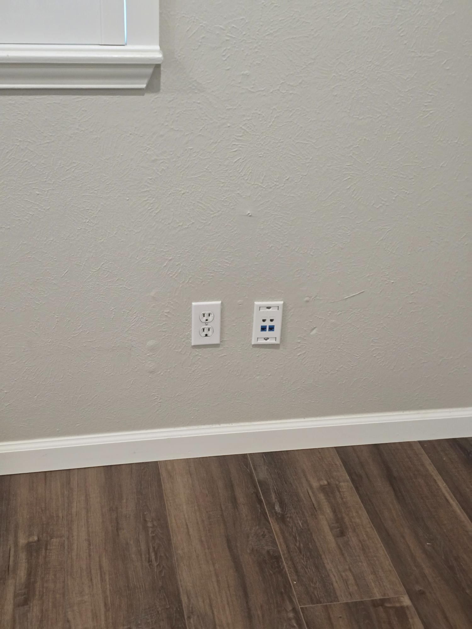 White electrical outlets and a blue-port data outlet are mounted on a gray wall above brown wood flooring.