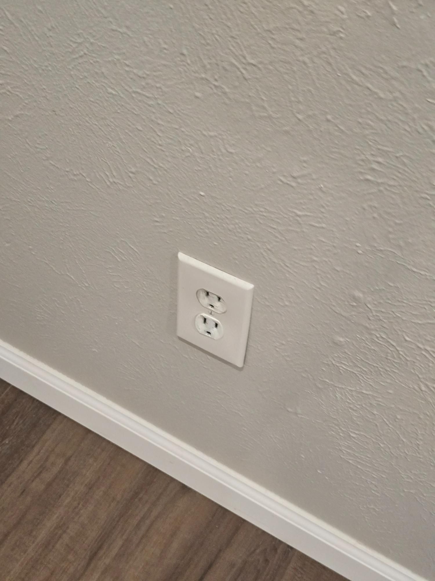 White electrical outlet on a textured light gray wall with wood-look flooring below.