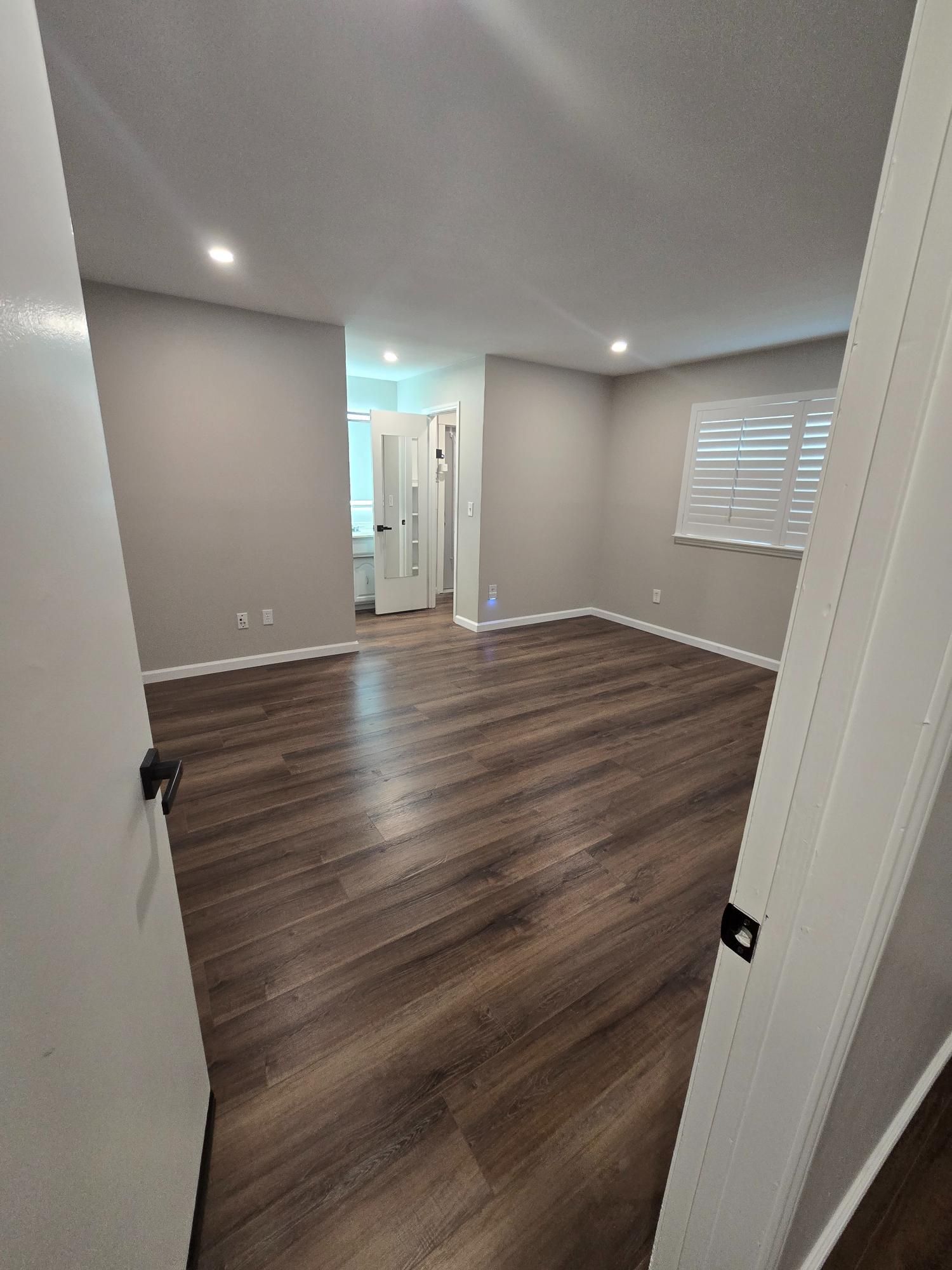 Empty room with wood floors and gray walls. Window with blinds, recessed lighting. Open door frames visible.