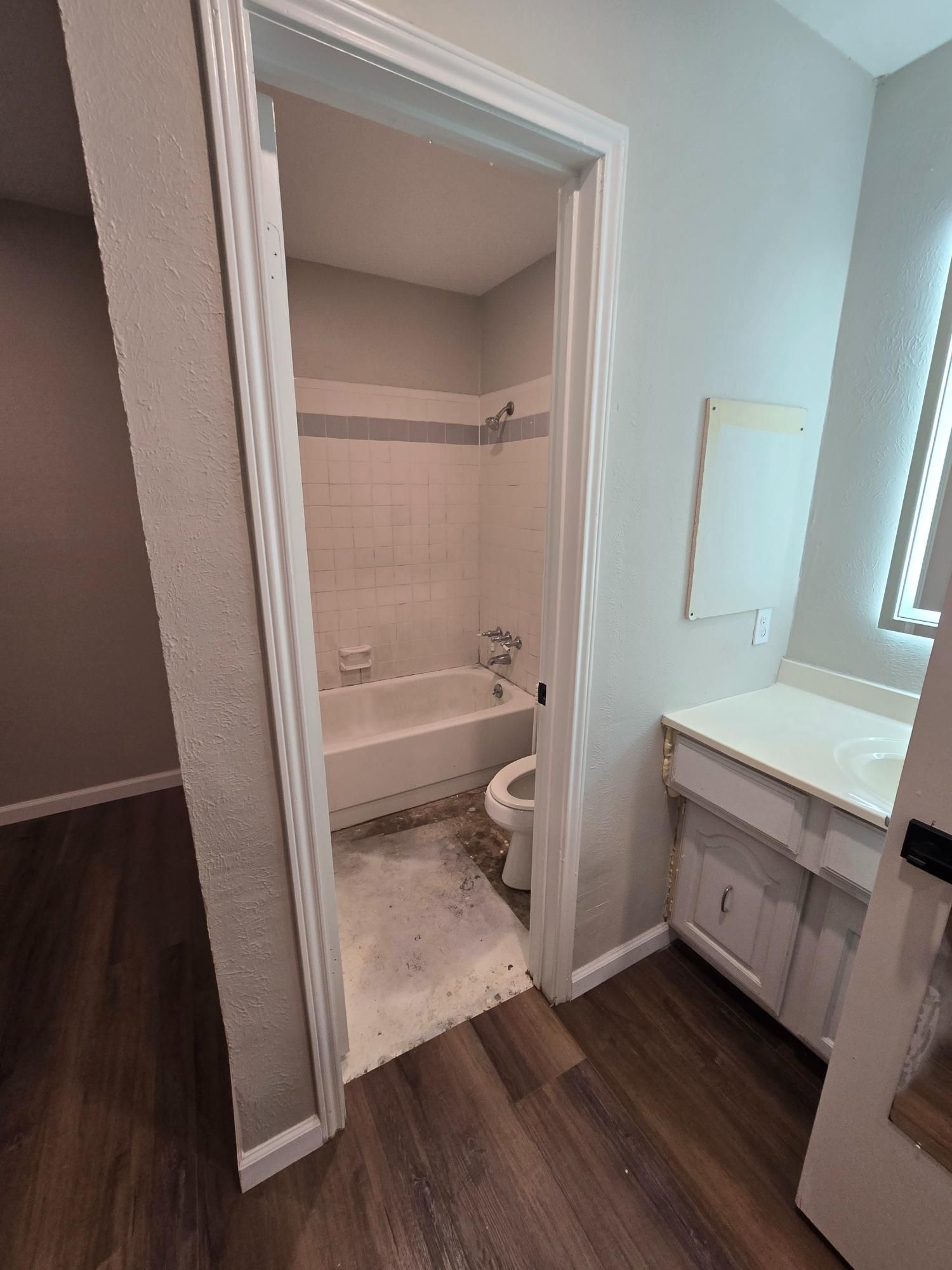 Bathroom with a white bathtub, toilet, vanity, and brown flooring, accessible through a doorway.