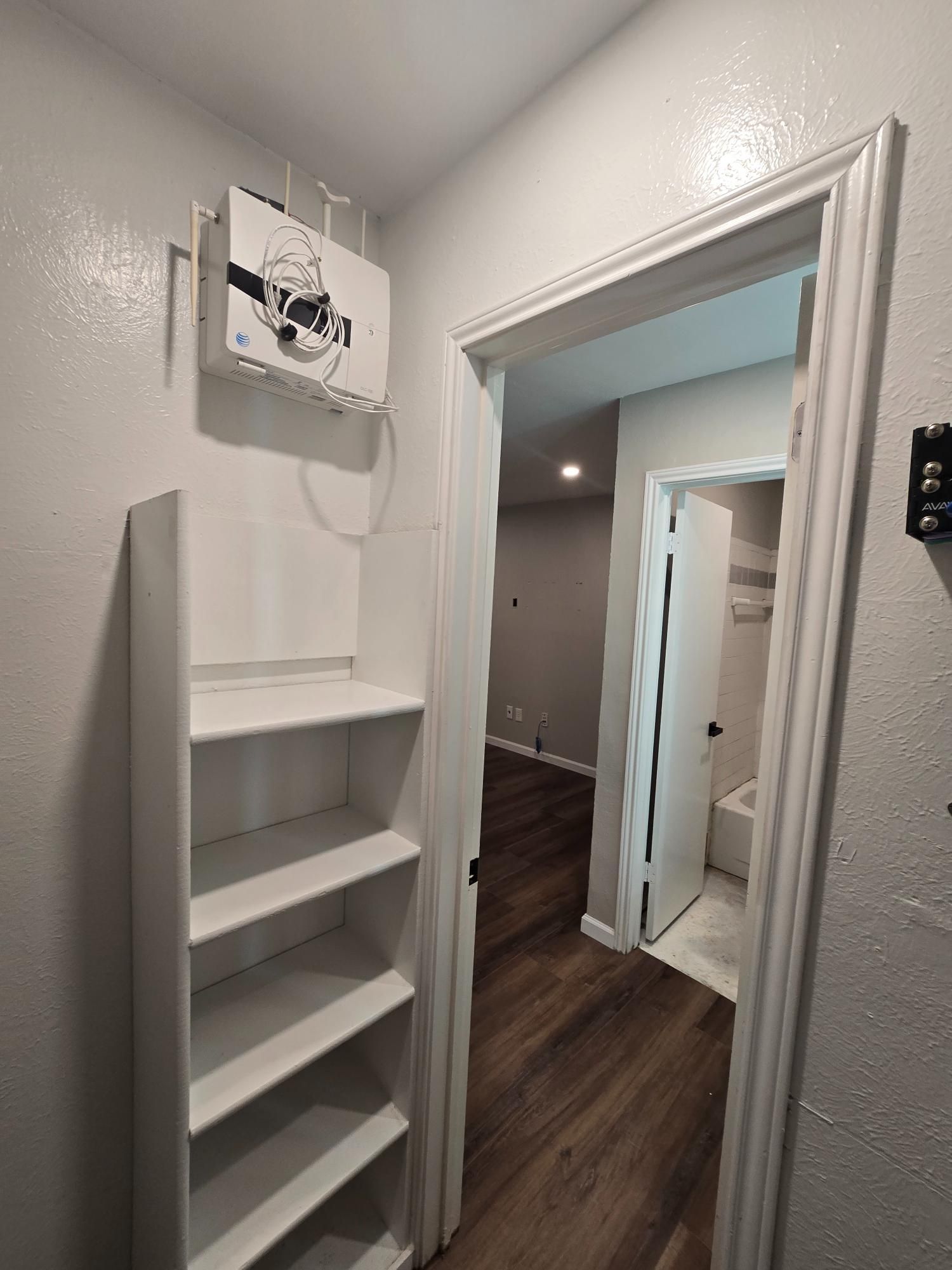 Narrow hallway with white shelves and doorway leading to a room with wood flooring and a bathroom.
