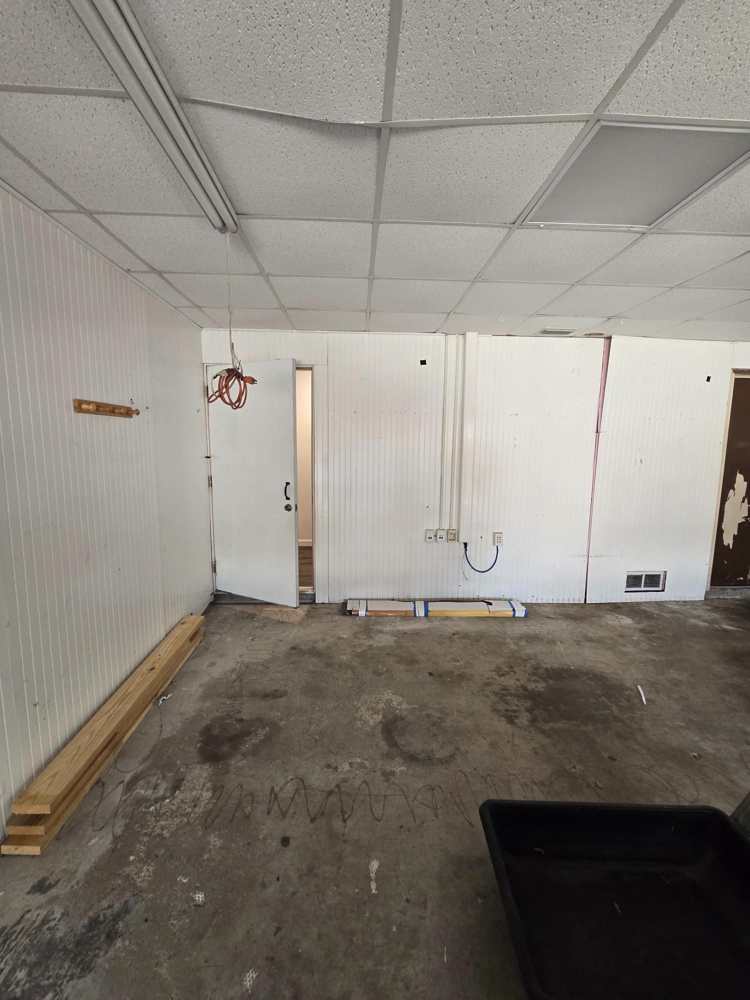 Empty room with damaged walls, ceiling tiles, and concrete floor. Boards and wires present.