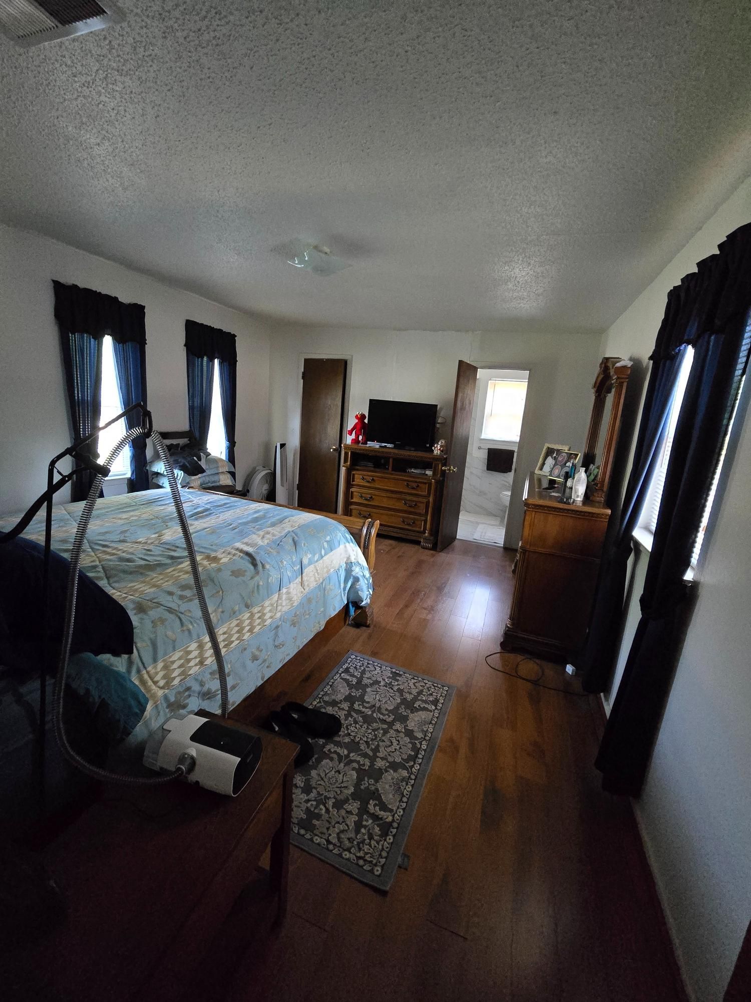 Bedroom with bed, dresser, TV, and open doorway to a bathroom; hardwood floor.