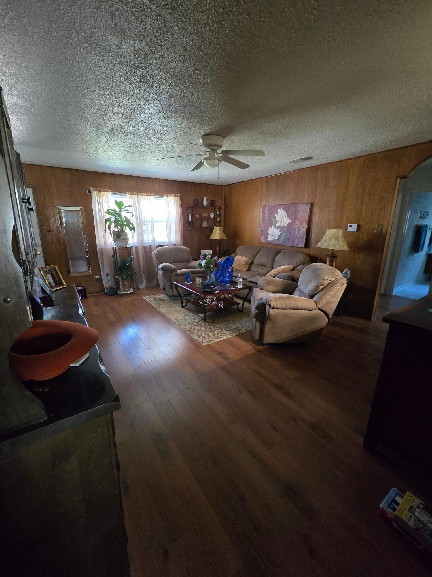 Living room with wood paneling, dark floors, couch, recliner, ceiling fan, and window.