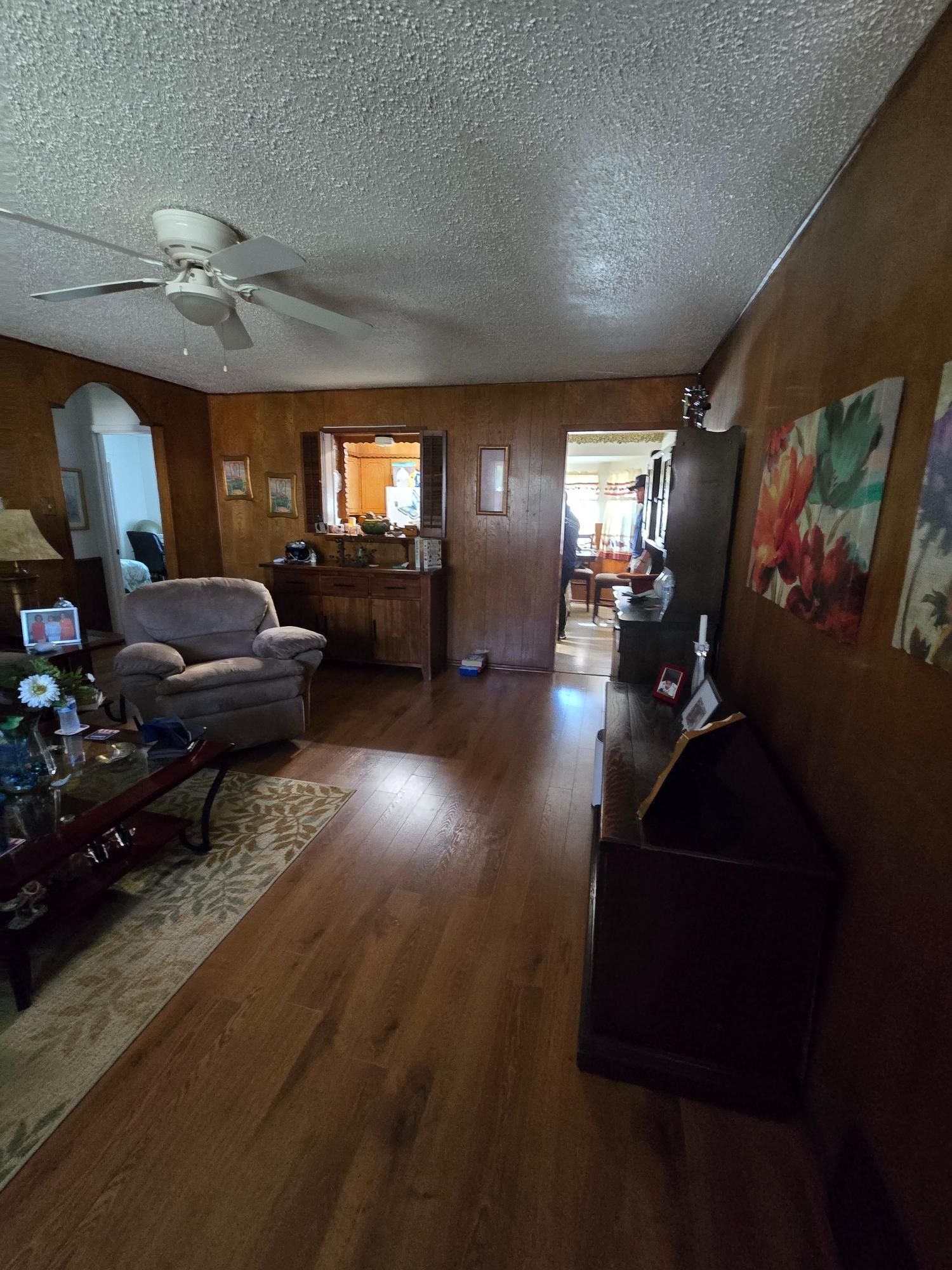Living room with wood paneling and brown flooring, ceiling fan. Armchair, rug, and art on the wall.