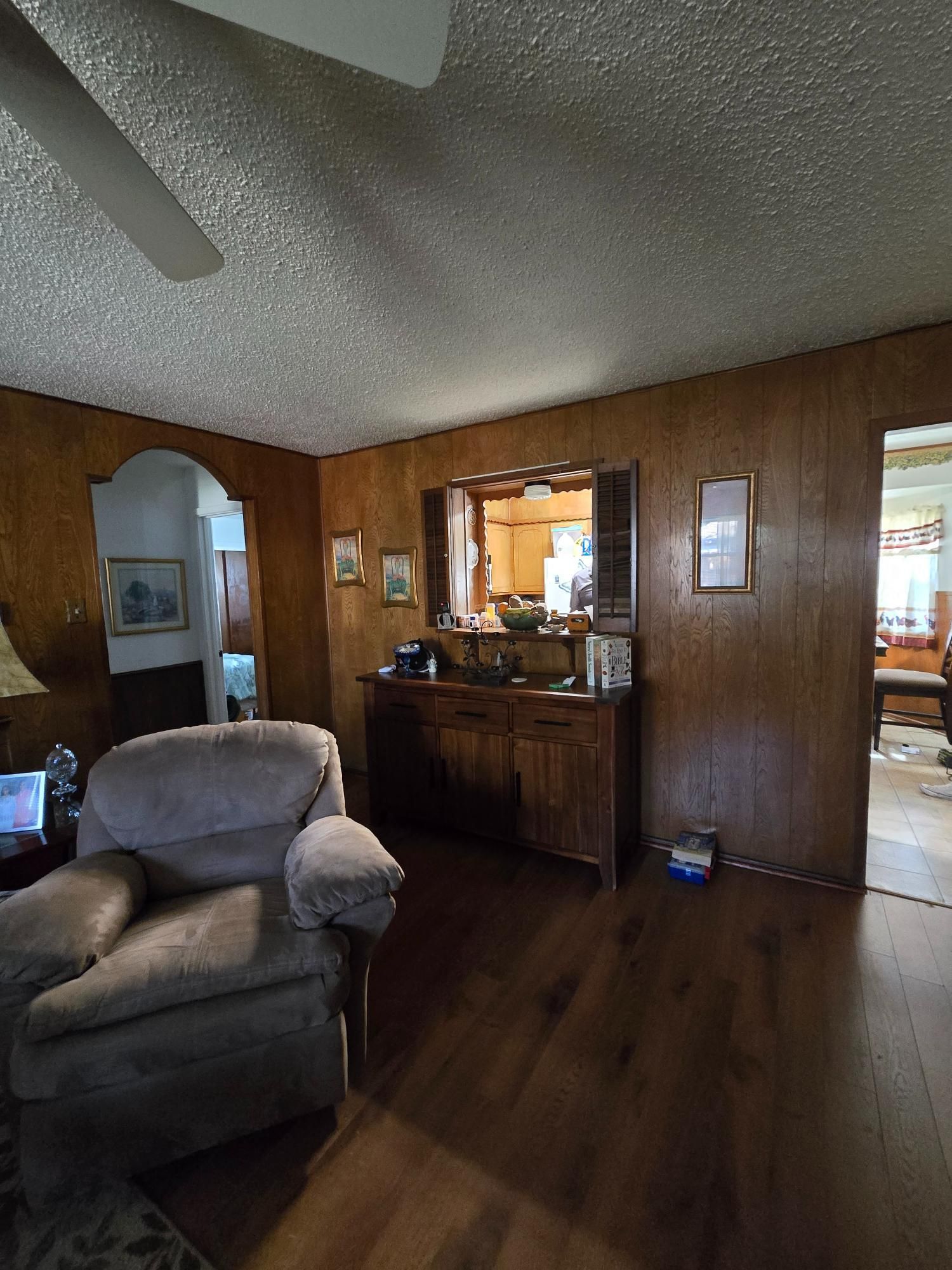 Cozy living room with wood paneling, armchair, and dark wooden buffet. Archway leads to another room.