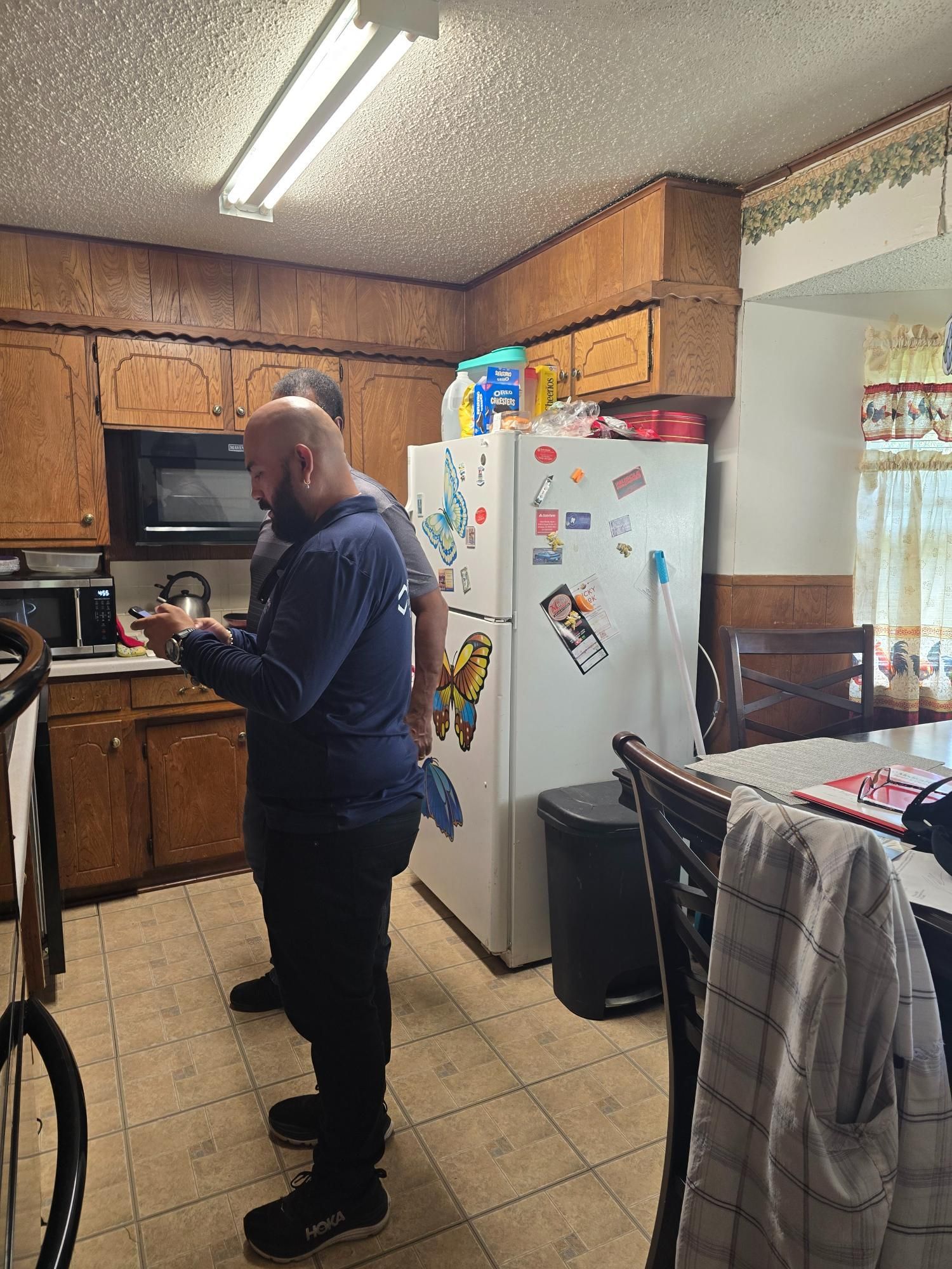 Two people in a kitchen; one looking at phone, refrigerator, microwave, wooden cabinets, dining table.