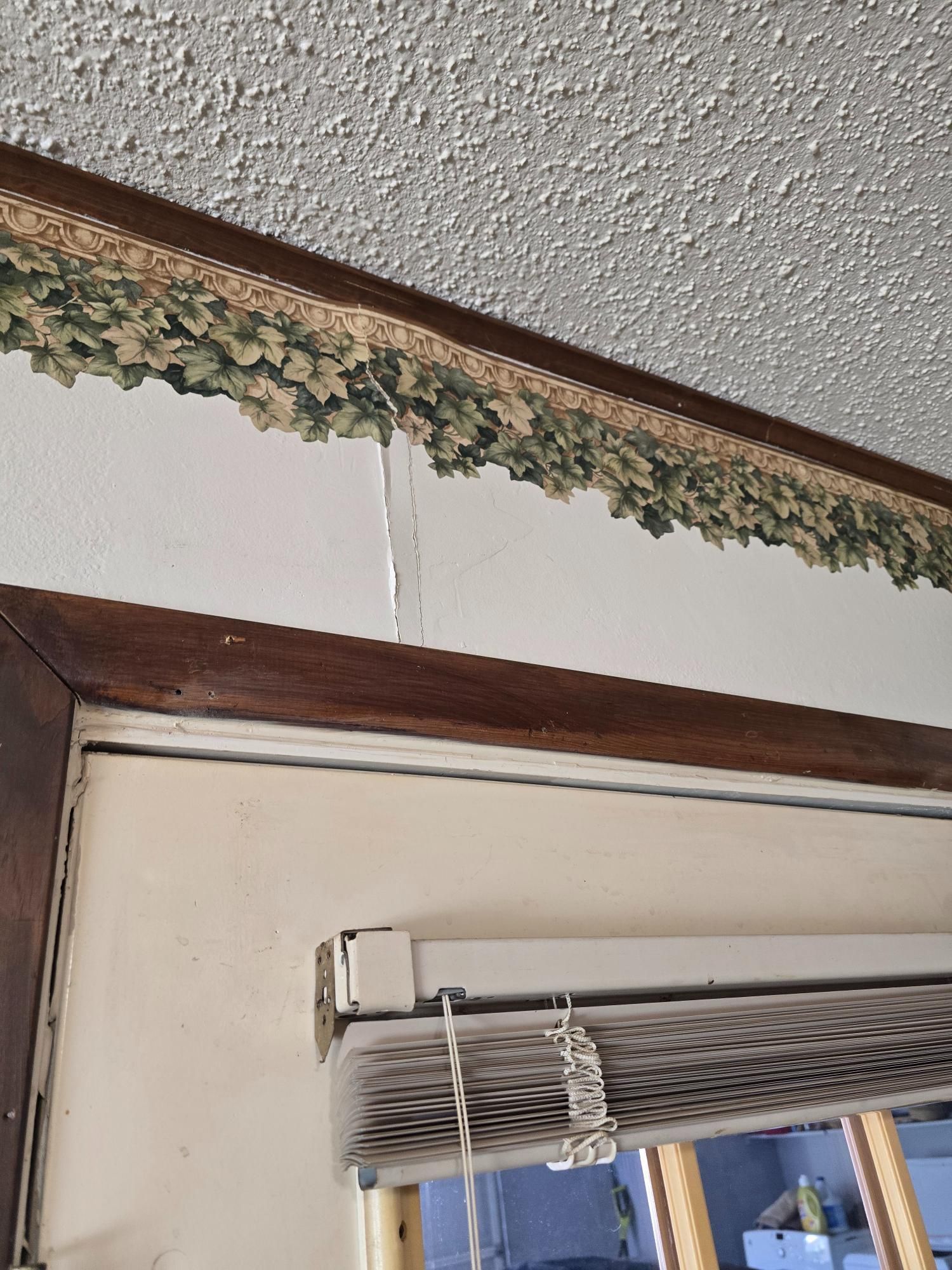 Brown trim above a window with a valance of leafy design; white wall, textured ceiling, and blinds.