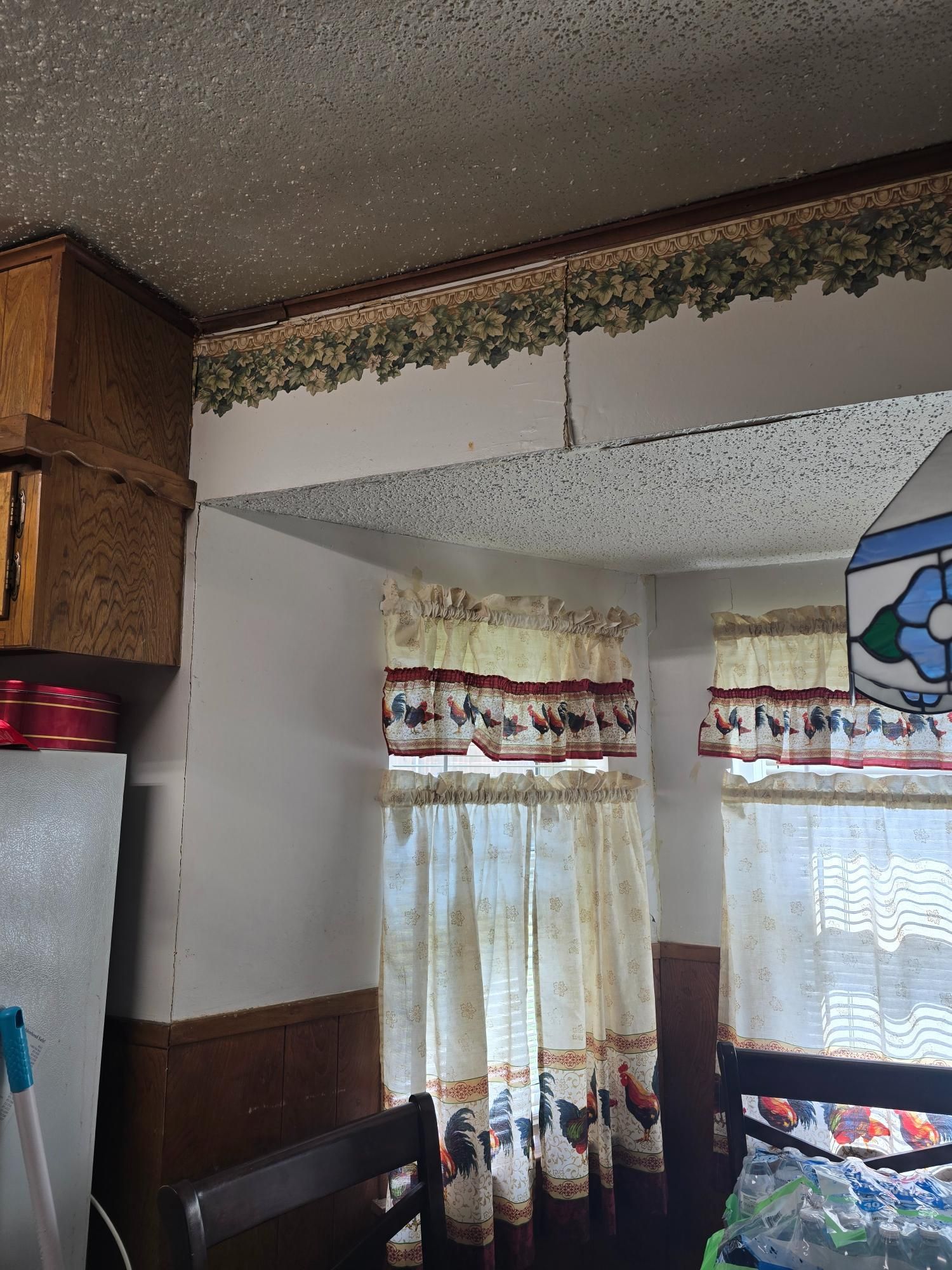 Kitchen interior: wooden cabinets, rooster curtains, wallpaper border, textured ceiling.