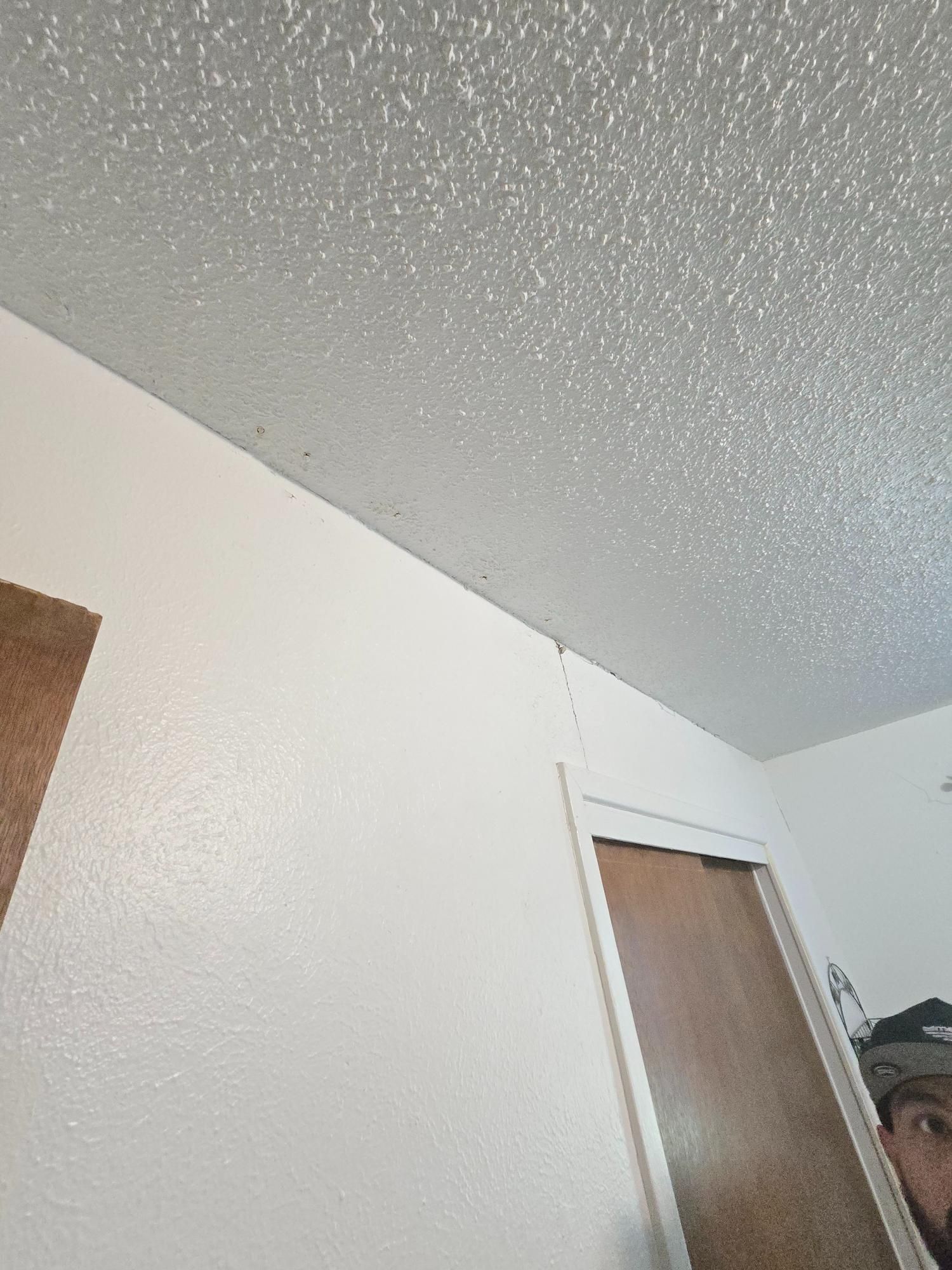 White walls and textured ceiling in a room, door on right, person peeking from behind the door.