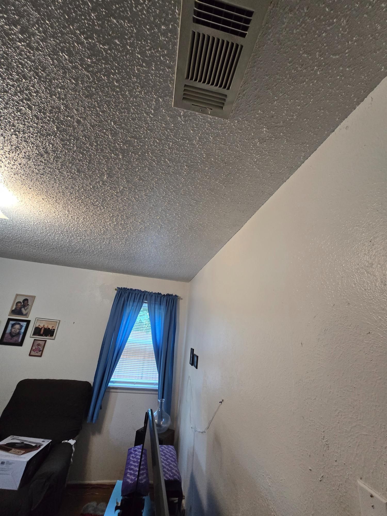 Room interior with textured ceiling, white walls, blue curtains, and a vent. Includes a chair and a window.