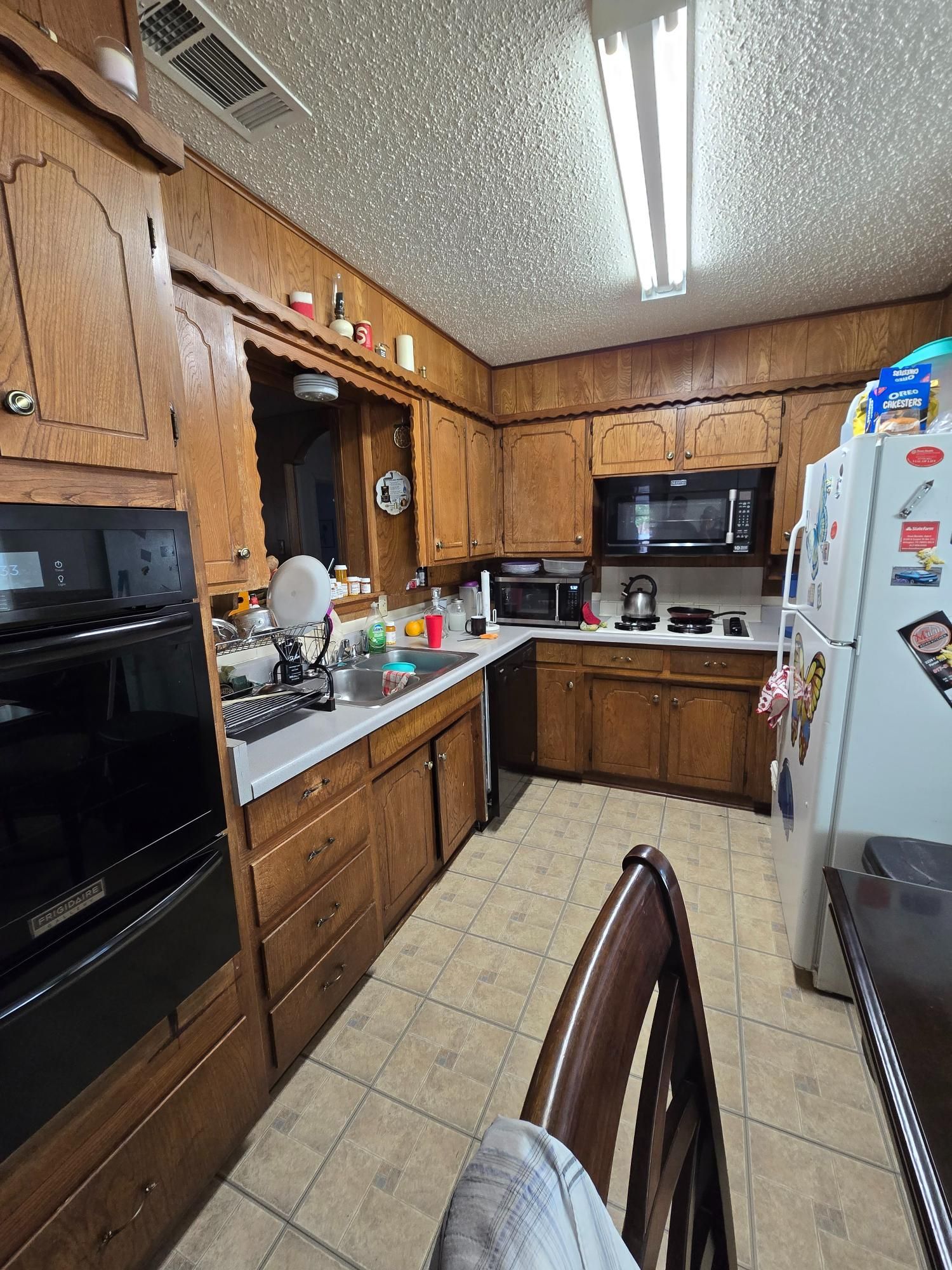 Kitchen with wood cabinets, appliances, and a white refrigerator; tile flooring and a ceiling light.