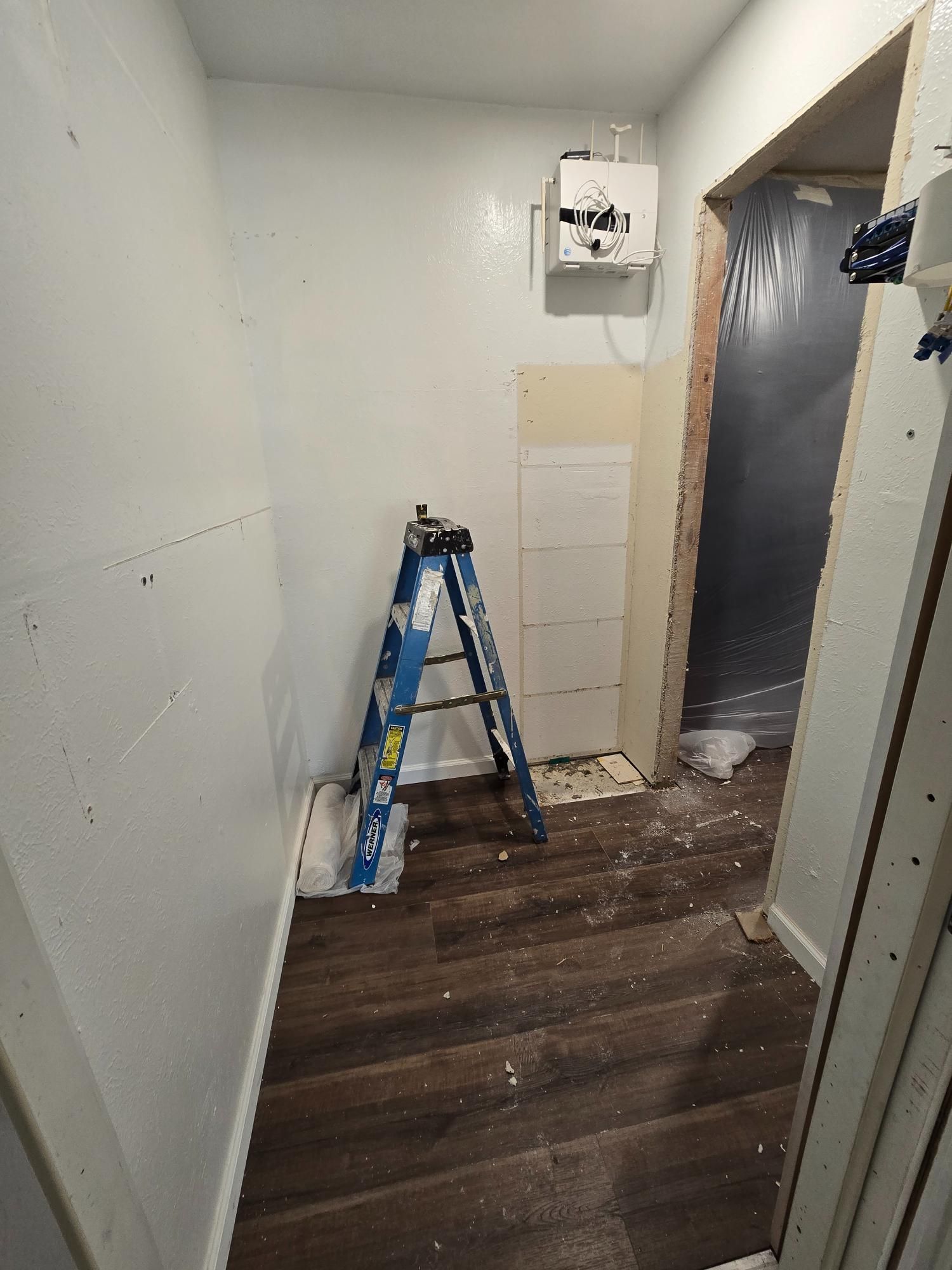 Hallway under construction: blue ladder, water heater, wood flooring, doorway covered in plastic, white walls.