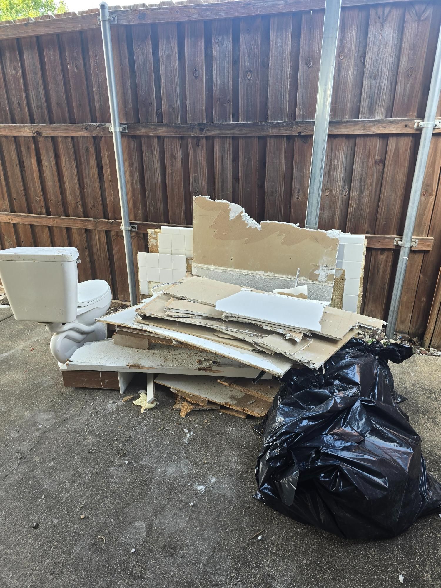 Pile of construction debris and toilet outside, next to a black trash bag and wooden fence.