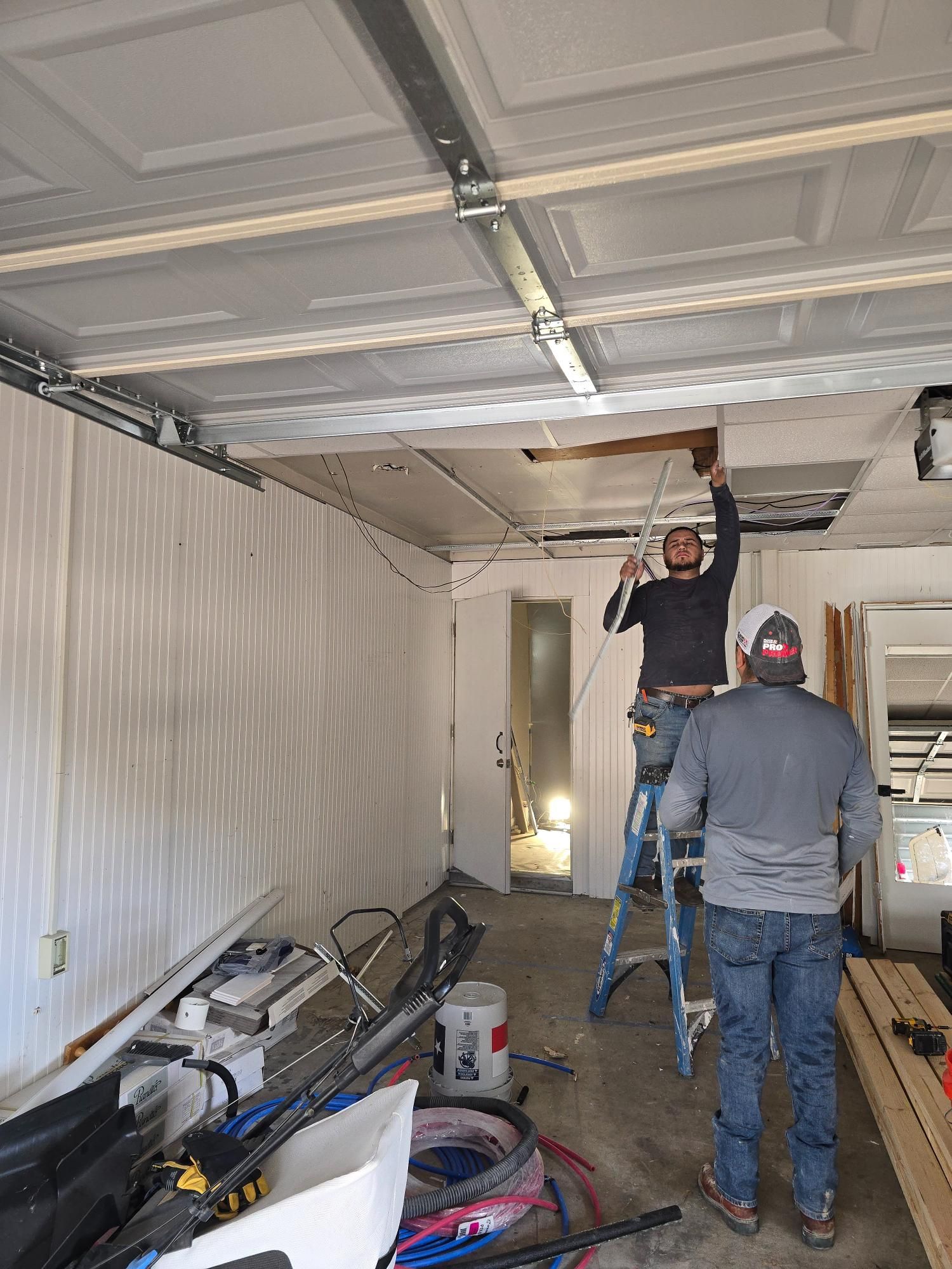 Two workers inside a garage, one on a ladder, working on the ceiling. Tools and debris on the floor.