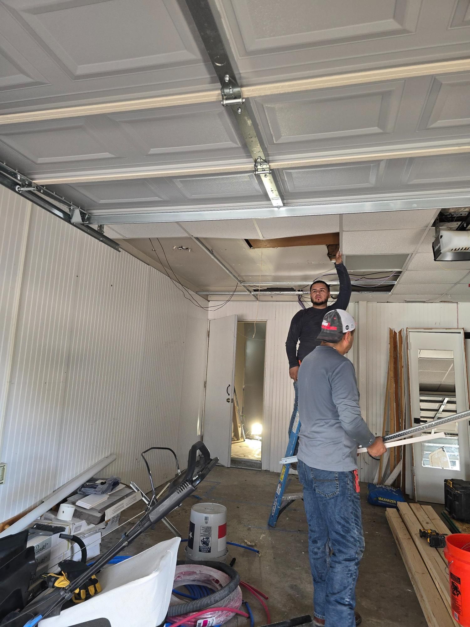 Men repairing a garage ceiling. One on a ladder, the other holding materials. White ceiling, debris on the floor.