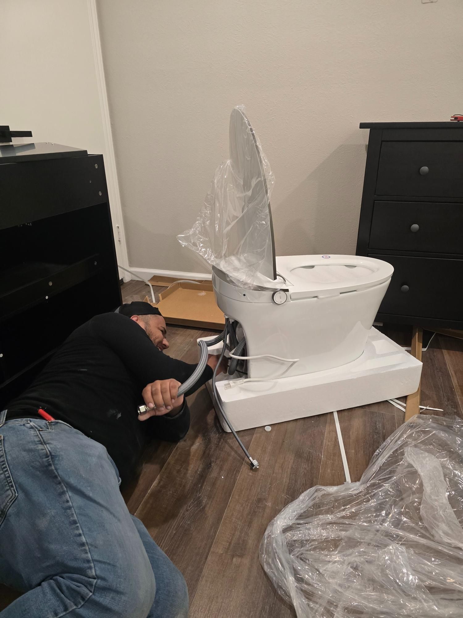 Person installing a white toilet in a room with wood flooring, kneeling and using a tool.
