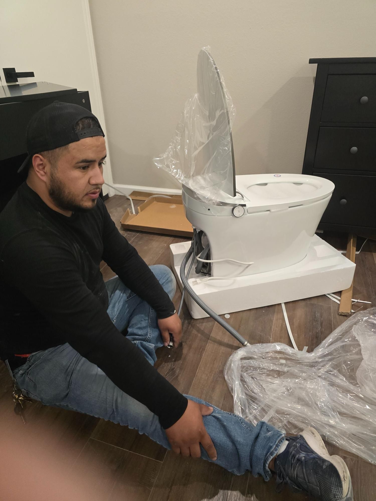 Man sits next to a new toilet on a cardboard base in a room. He looks at his leg.
