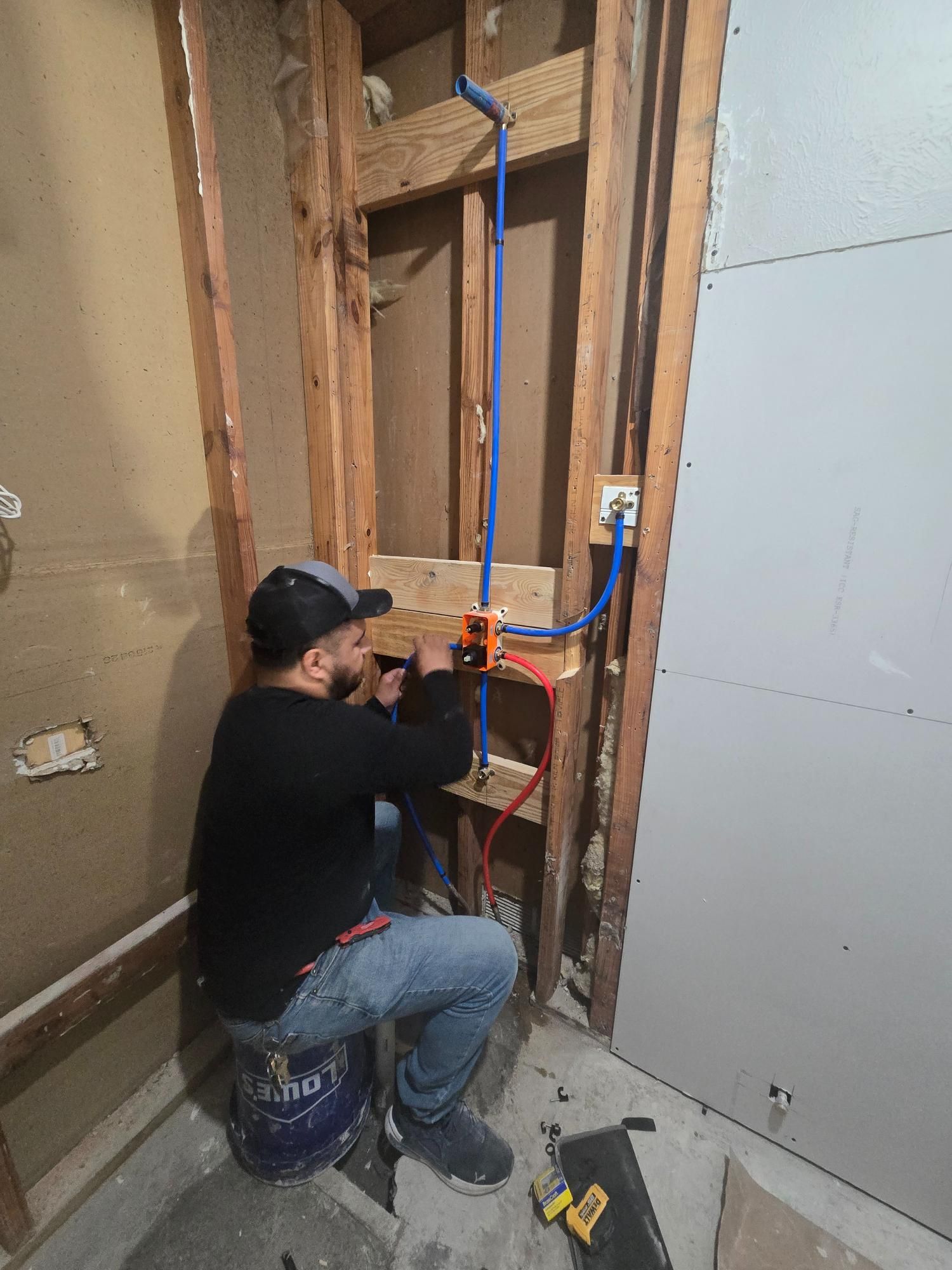 Plumber working on water pipes inside a wall, using tools. Blue and red PEX tubing, wood framing.