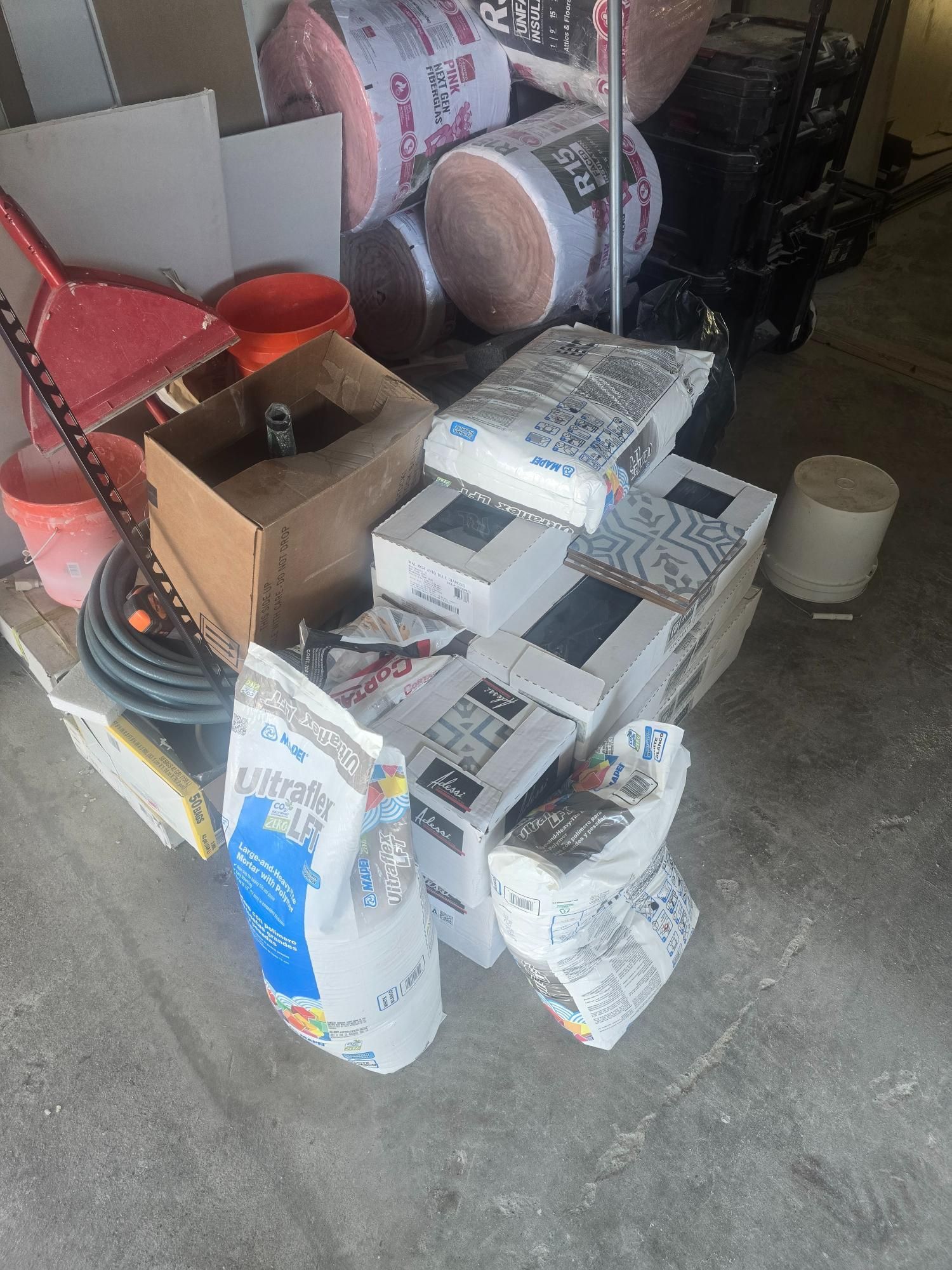 Pile of construction materials, including bags, boxes, insulation rolls, and buckets, on a concrete floor.