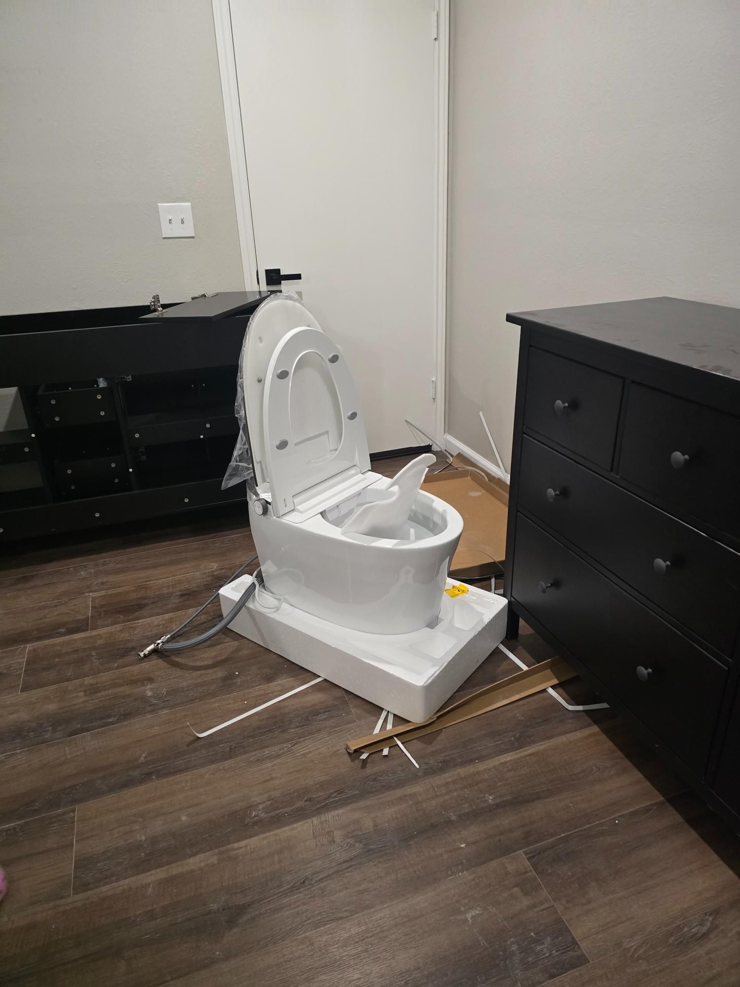 White, new toilet sitting on a white base in a room. Furniture and cardboard boxes are nearby.