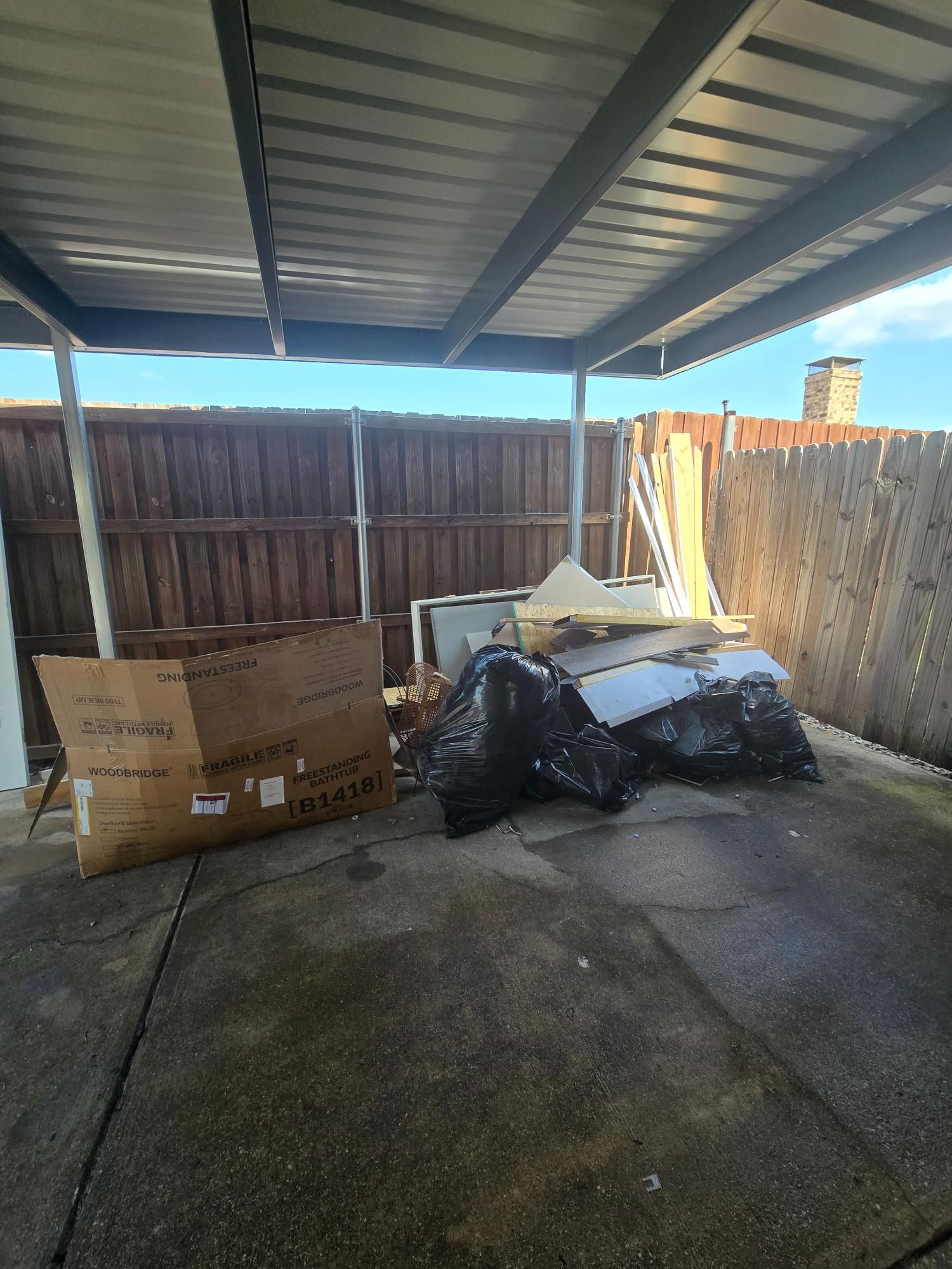A carport with cardboard boxes and black trash bags. A wooden fence is in the background.
