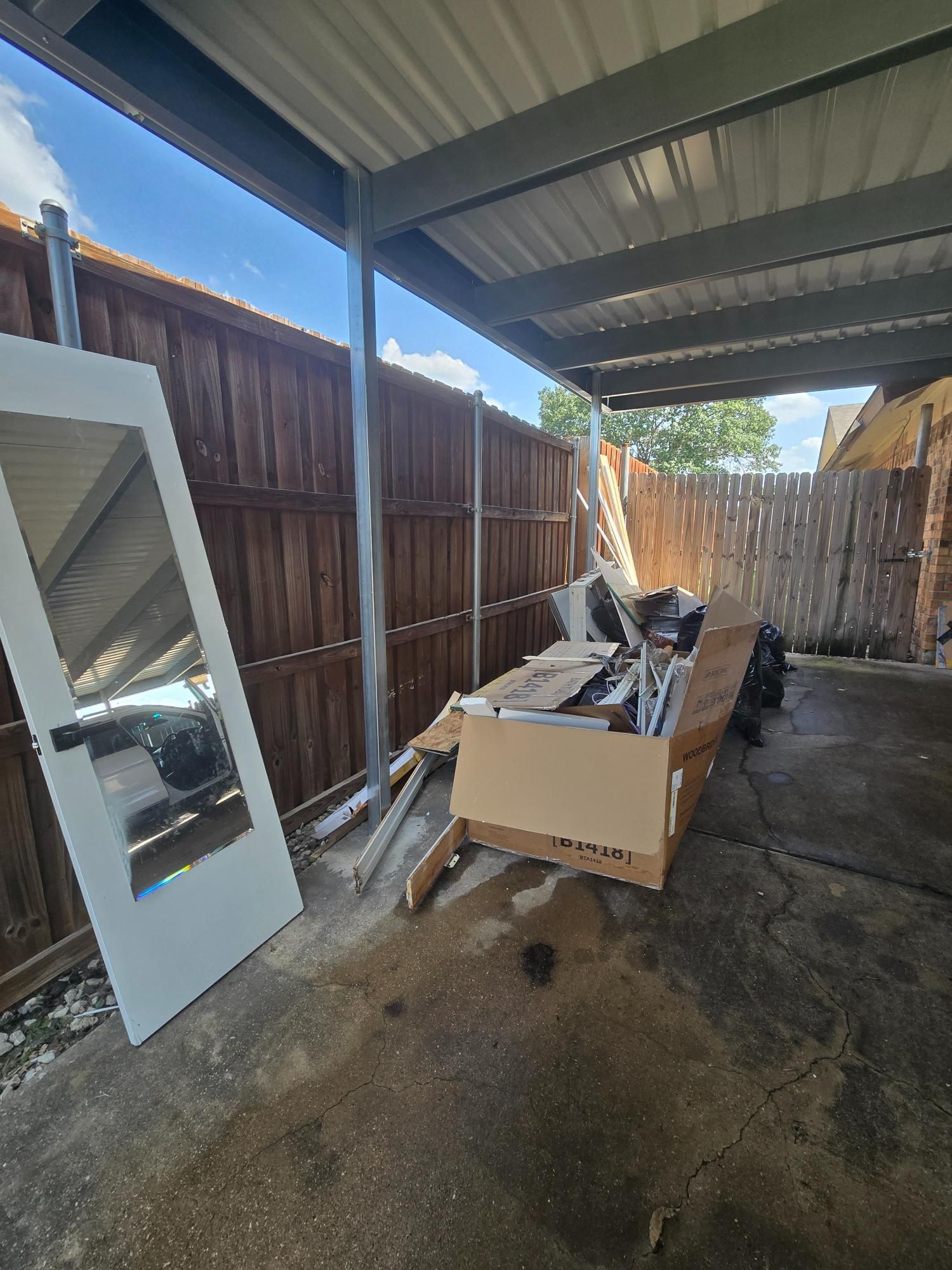 A doorway, debris, and boxes sit under a metal awning next to a wooden fence.
