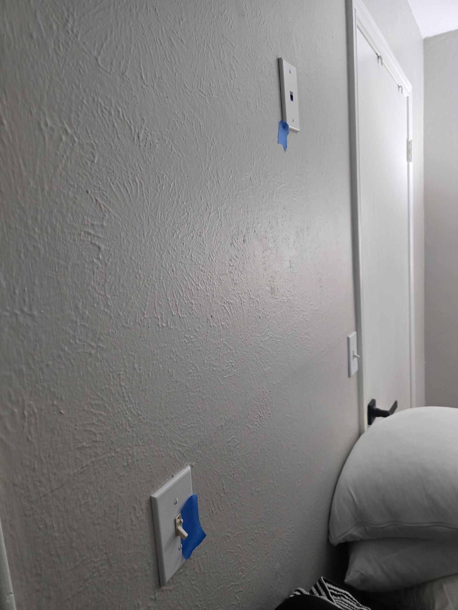 White wall with two electrical outlets covered in blue tape. A closed white door is nearby.