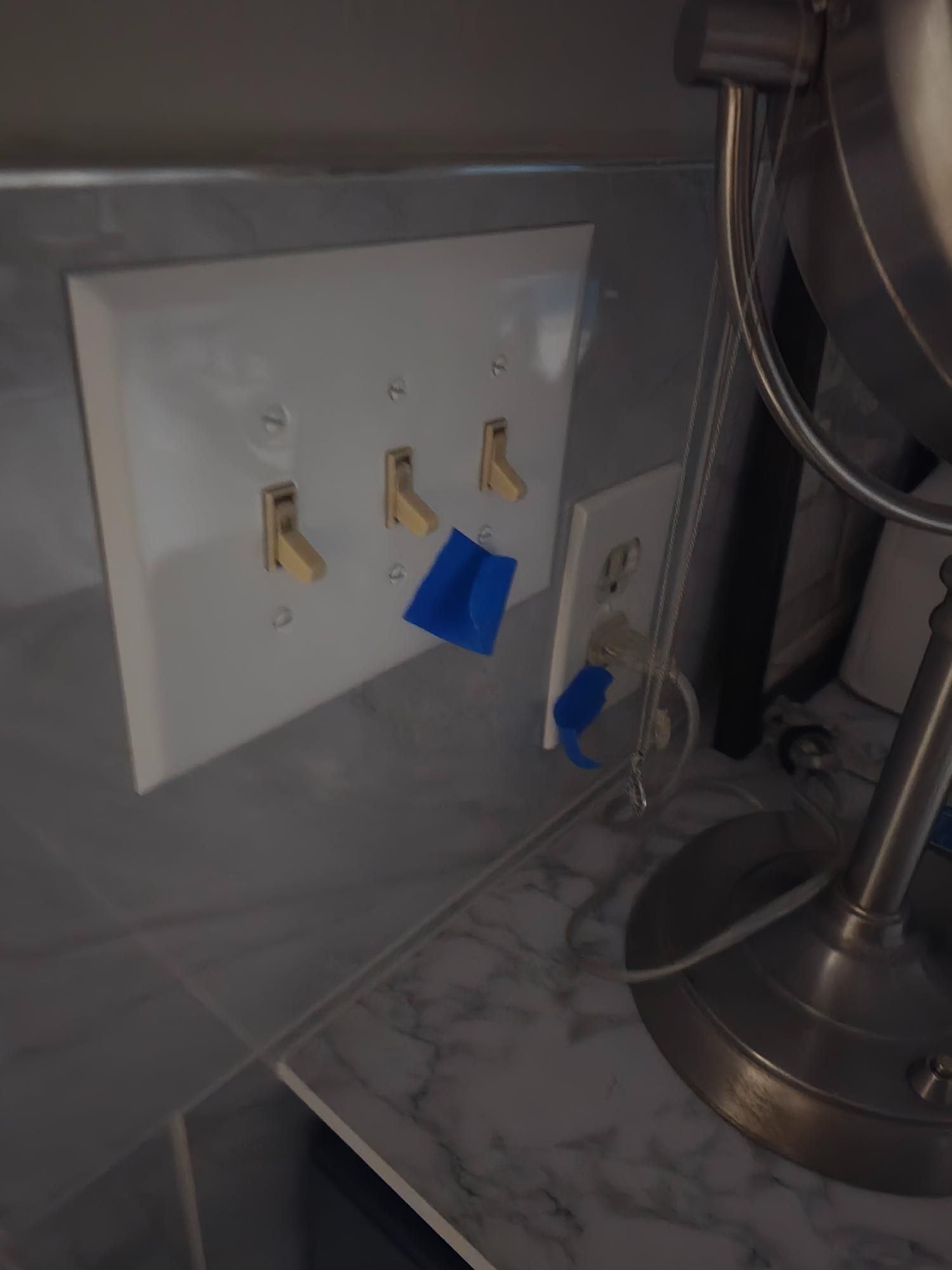 White switch plate with four light switches, blue tape, and an electrical outlet, beside a mirror.