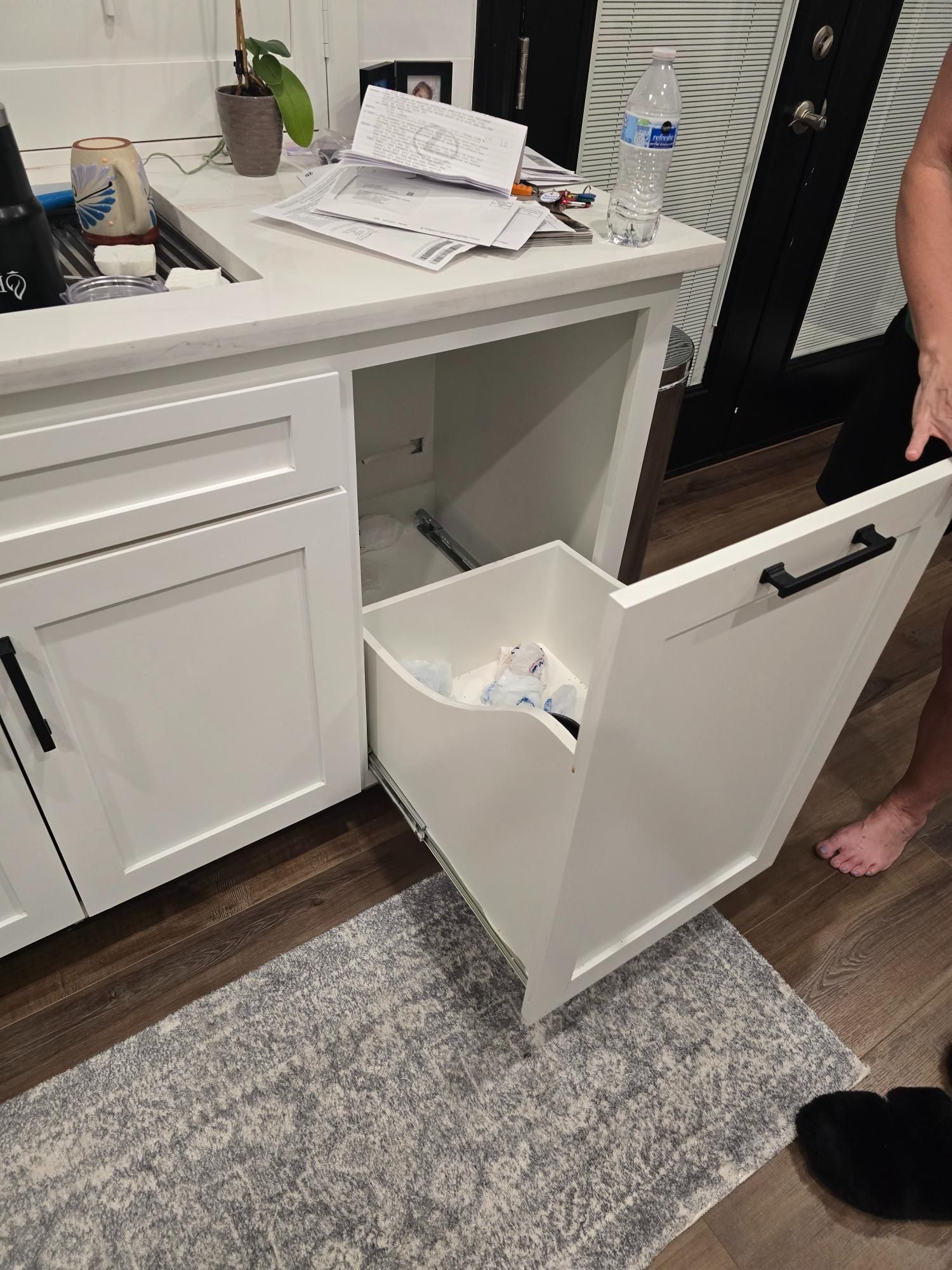White kitchen cabinet with pull-out trash bin open, revealing trash. Black handles.