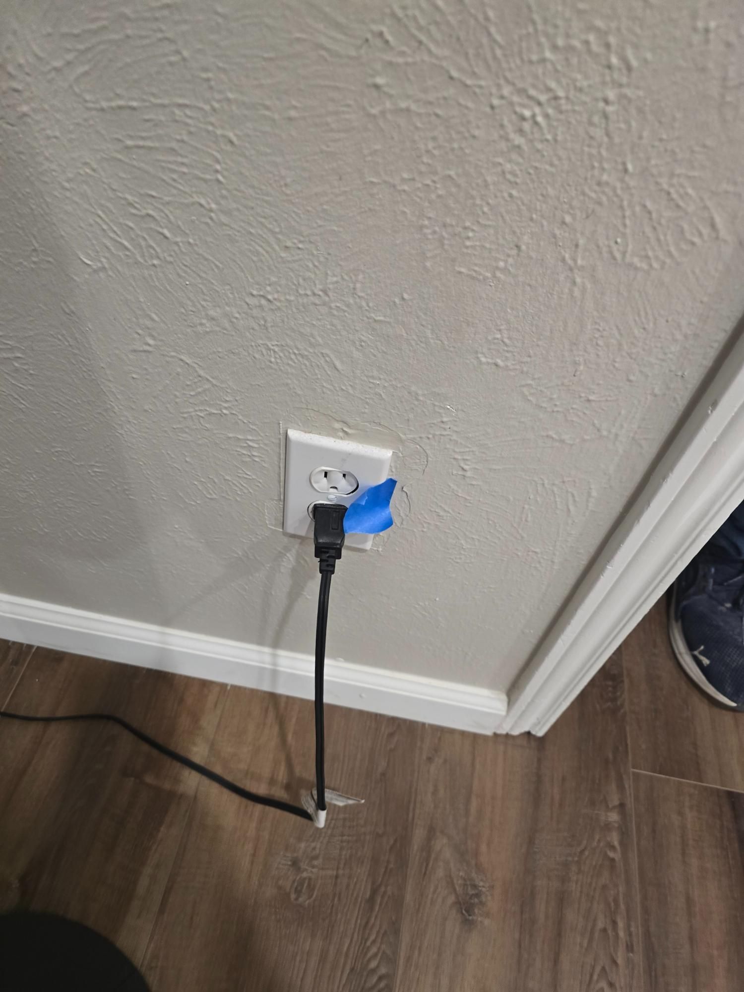 Power cord plugged into an electrical outlet on a beige wall, held in place with blue tape, on a wood floor.