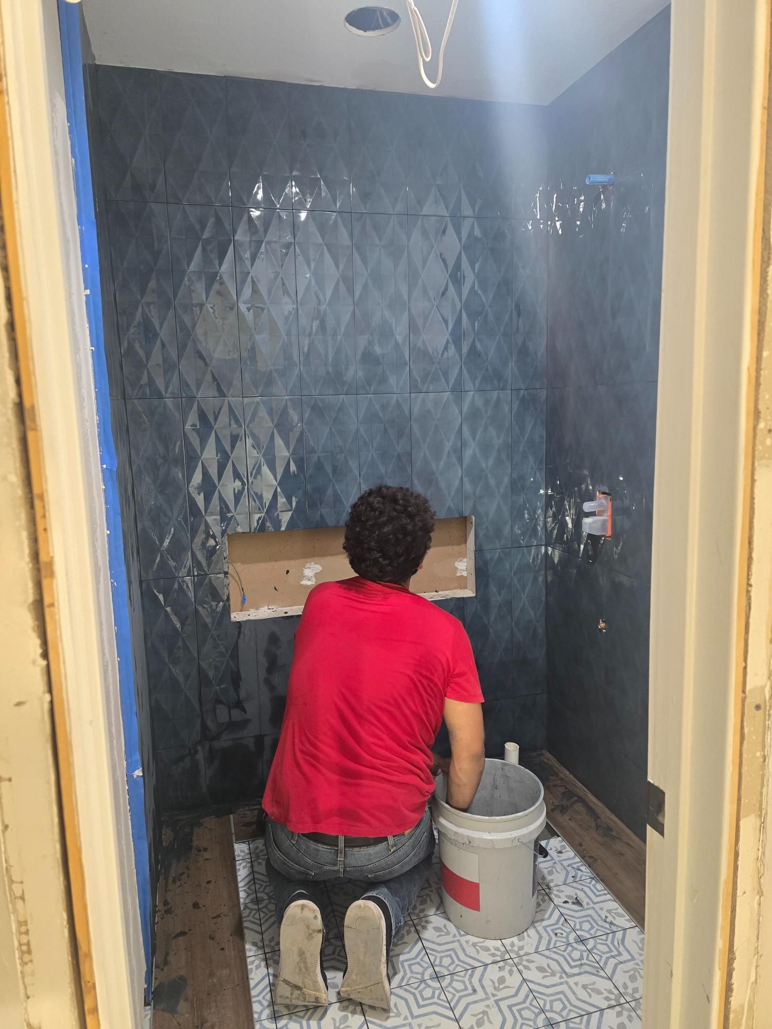 Person installing tiles in a blue-tiled shower, kneeling with bucket and tools.
