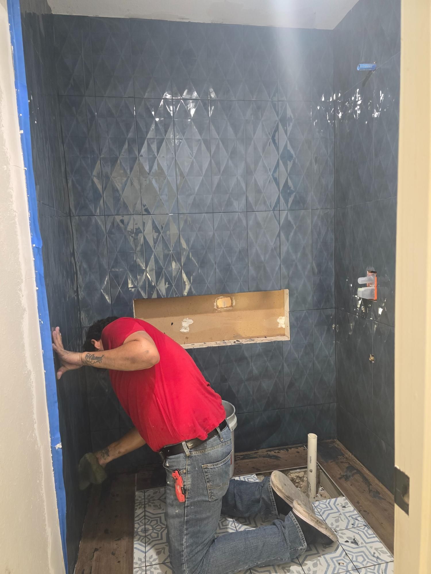 Man tiling a blue diamond-patterned shower wall, kneeling on a patterned mat.