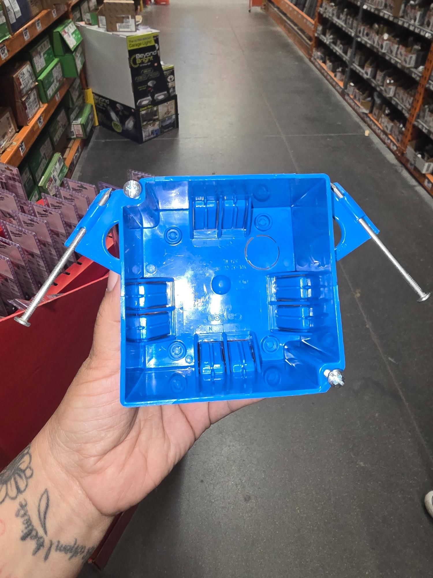 Blue electrical box with adjustable mounting arms held in a person's hand, inside a store.