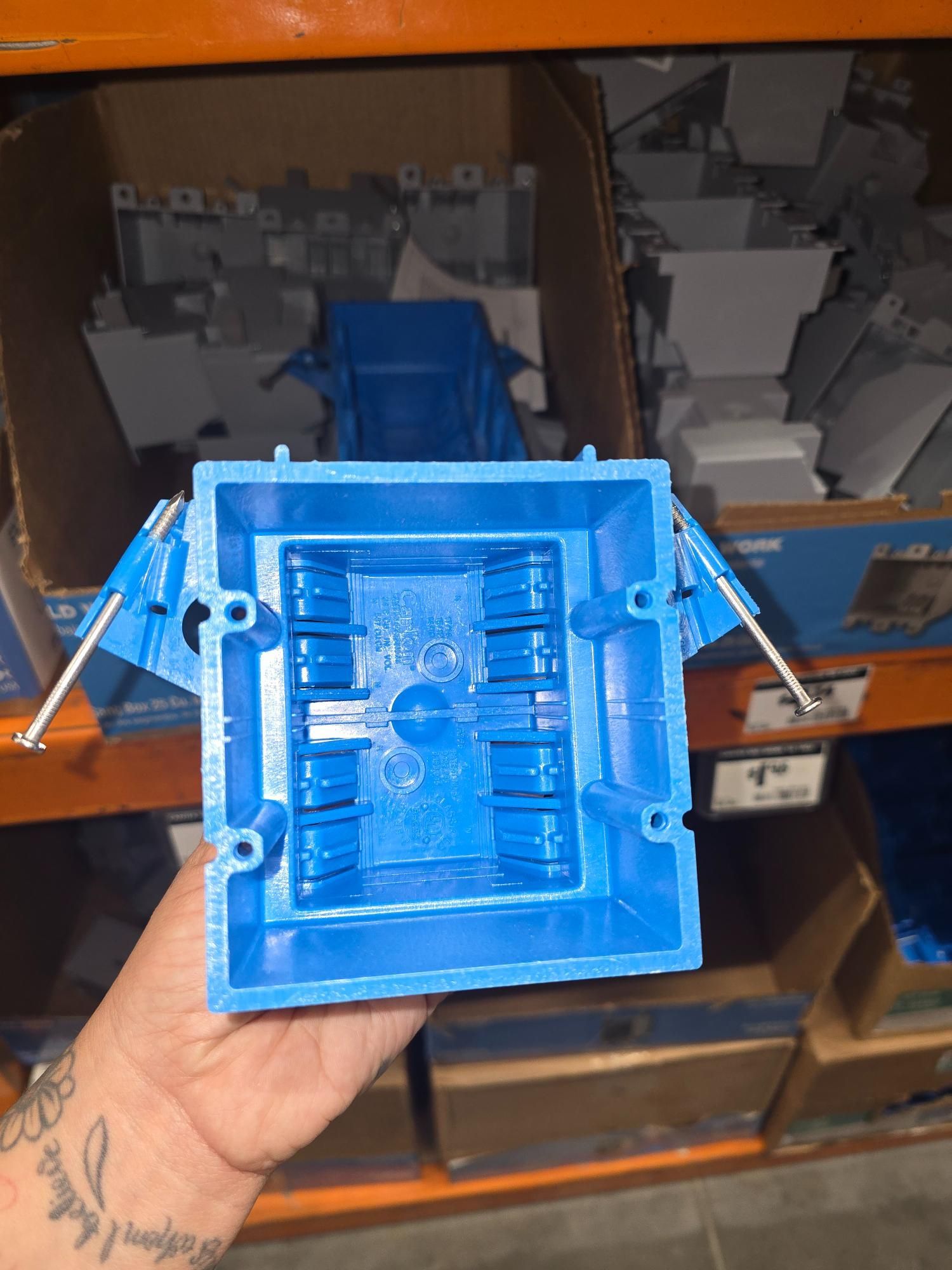 Blue electrical box held by a hand in a store, with screws extending out, surrounded by other boxes.