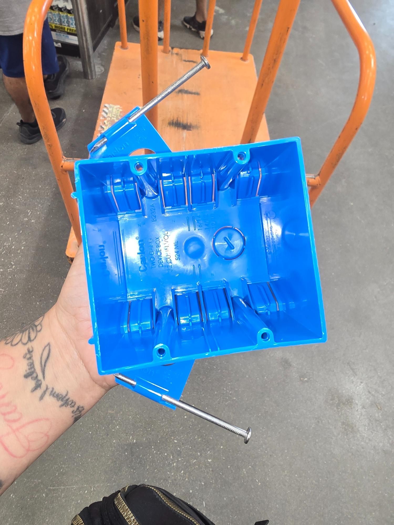 A blue electrical box held in hand, metal rods attached, orange cart in background.