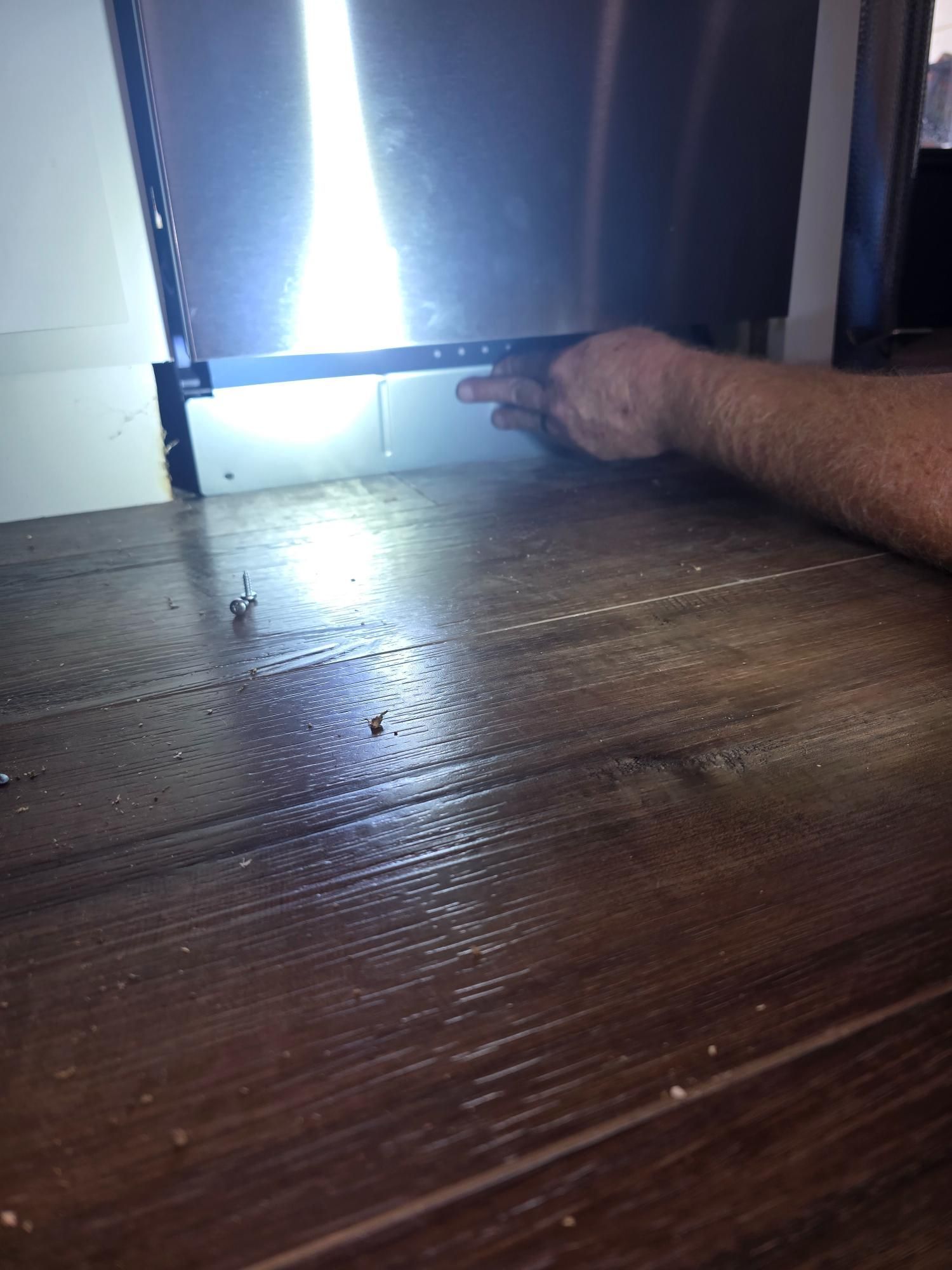 Person's hand pointing at a white panel beneath a stainless steel dishwasher, resting on wood flooring.