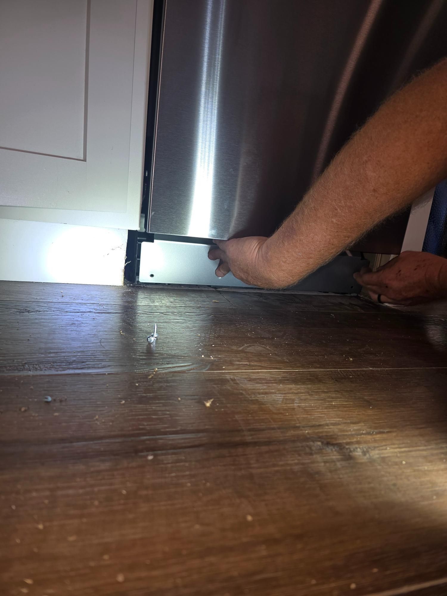 Person adjusts refrigerator foot, near white door and wood floor.