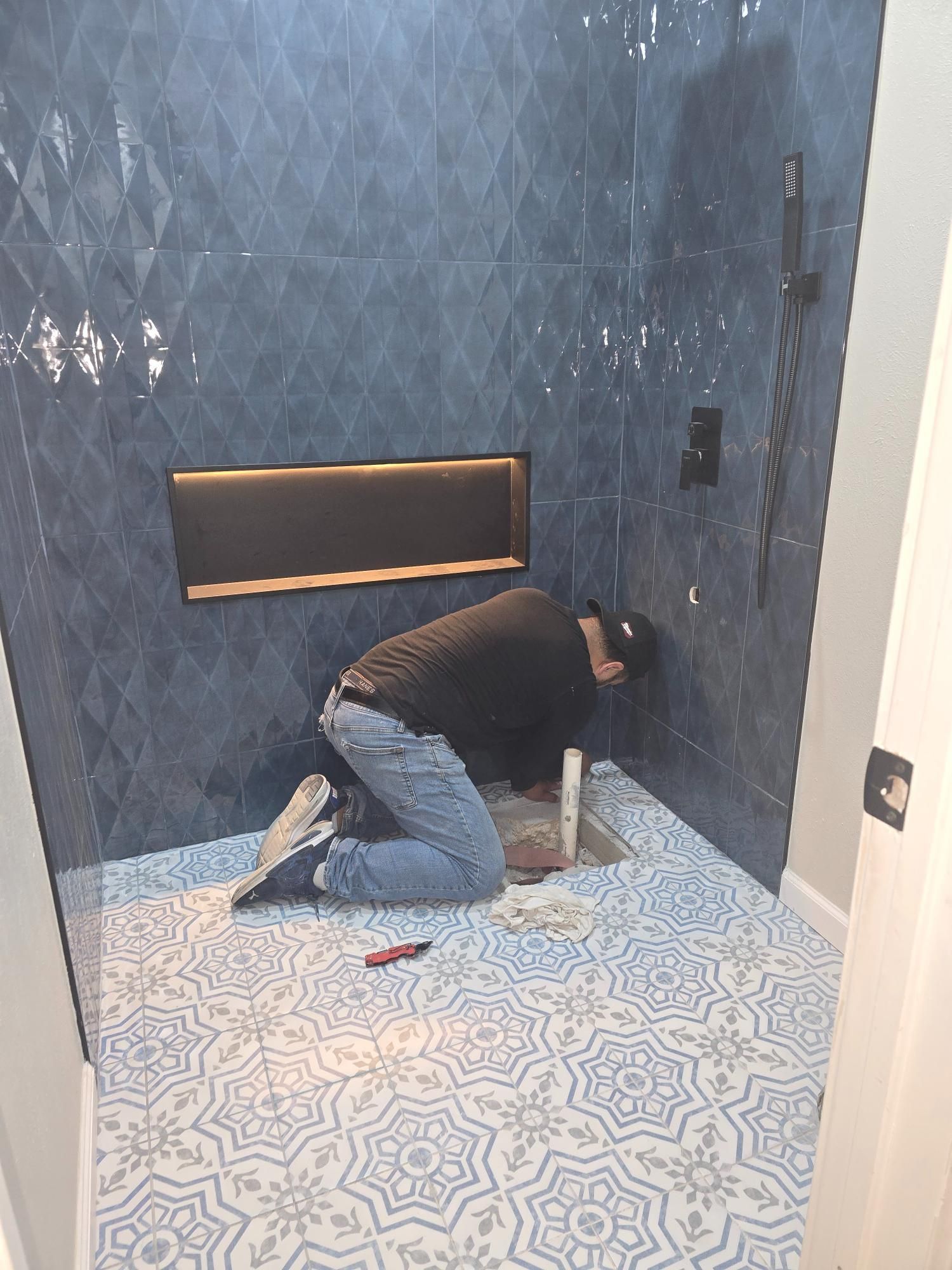 A person installing a tile shower. They are kneeling in the tiled space. The walls are blue and the floor has a pattern.